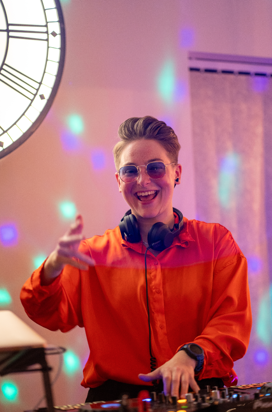 A person wearing sunglasses and headphones, smiling and pointing, standing behind a DJ mixing console at a party with colorful lights and a large clock on the wall in the background.