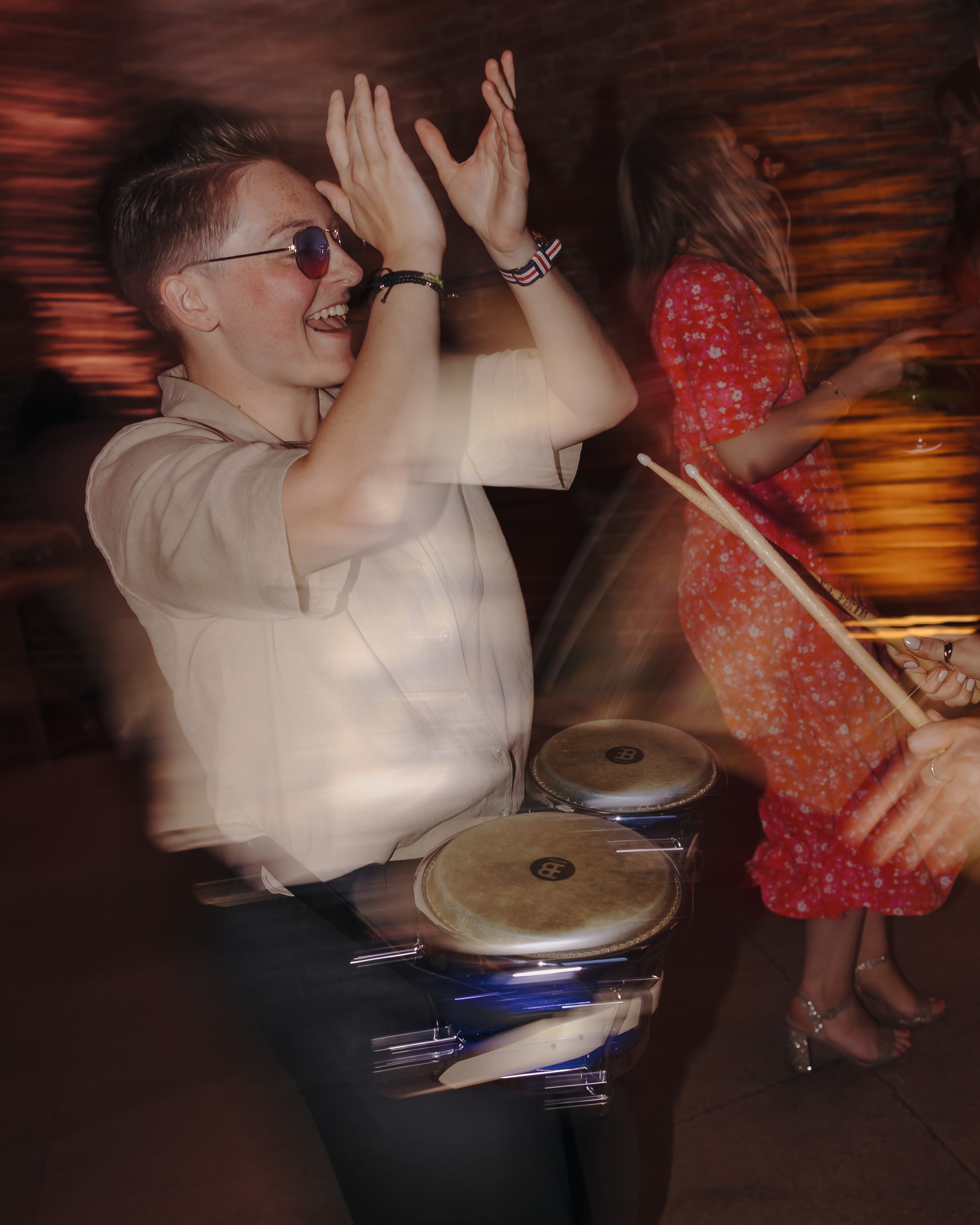 Frankie Santana dancing and playing bongos at a party, with a woman in a red dress holding drumsticks and playing the bongos too
