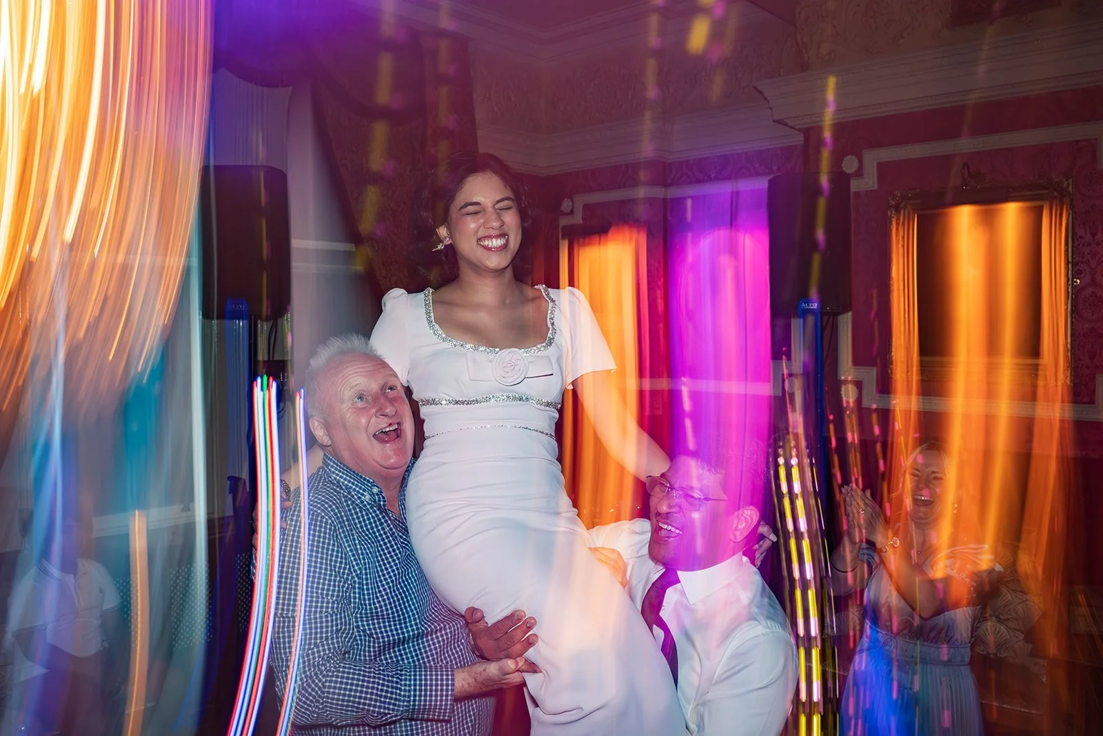 People celebrating at a party with colorful lights, including a woman being lifted by two men, all smiling and joyful.