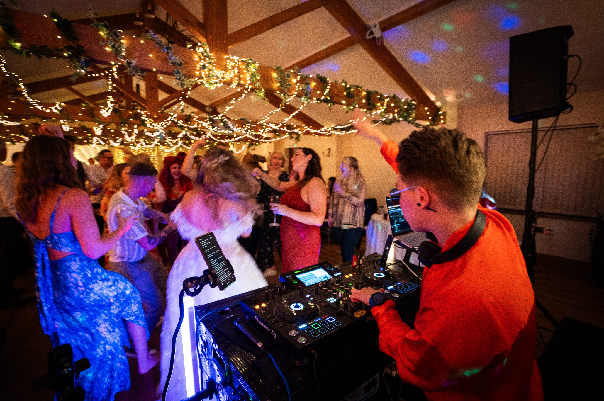 People dancing at a party or celebration with string lights and decorations overhead, a female DJ, Frankie Santana, playing music in the foreground.