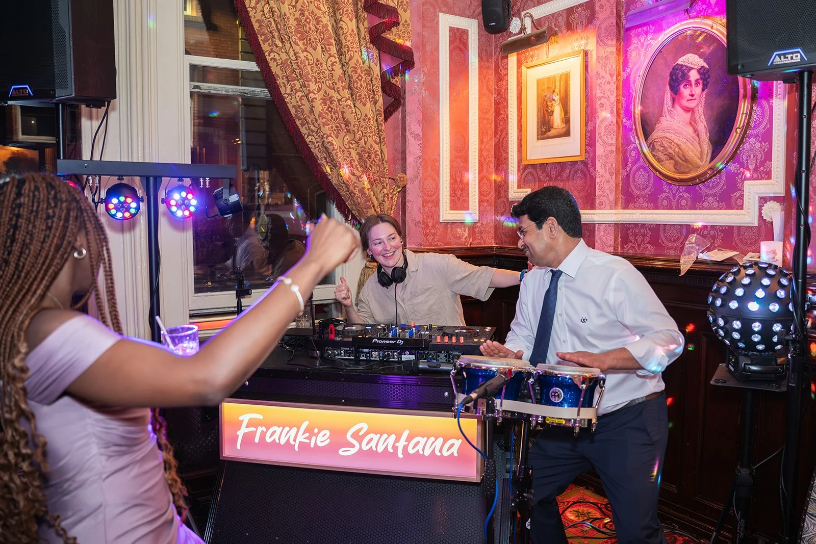 DJ woman with headphones and a customer playing bongo drums at a party with colorful lighting and vintage decor. Including framed portraits on pink wallpaper, and a woman dancing in the foreground