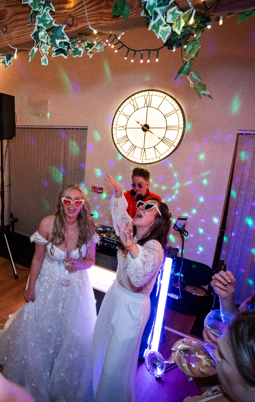 People at a party wearing retro sunglasses, dancing and enjoying drinks under colorful party lights with a large clock on the wall and decorative string lights and greenery hanging from the ceiling