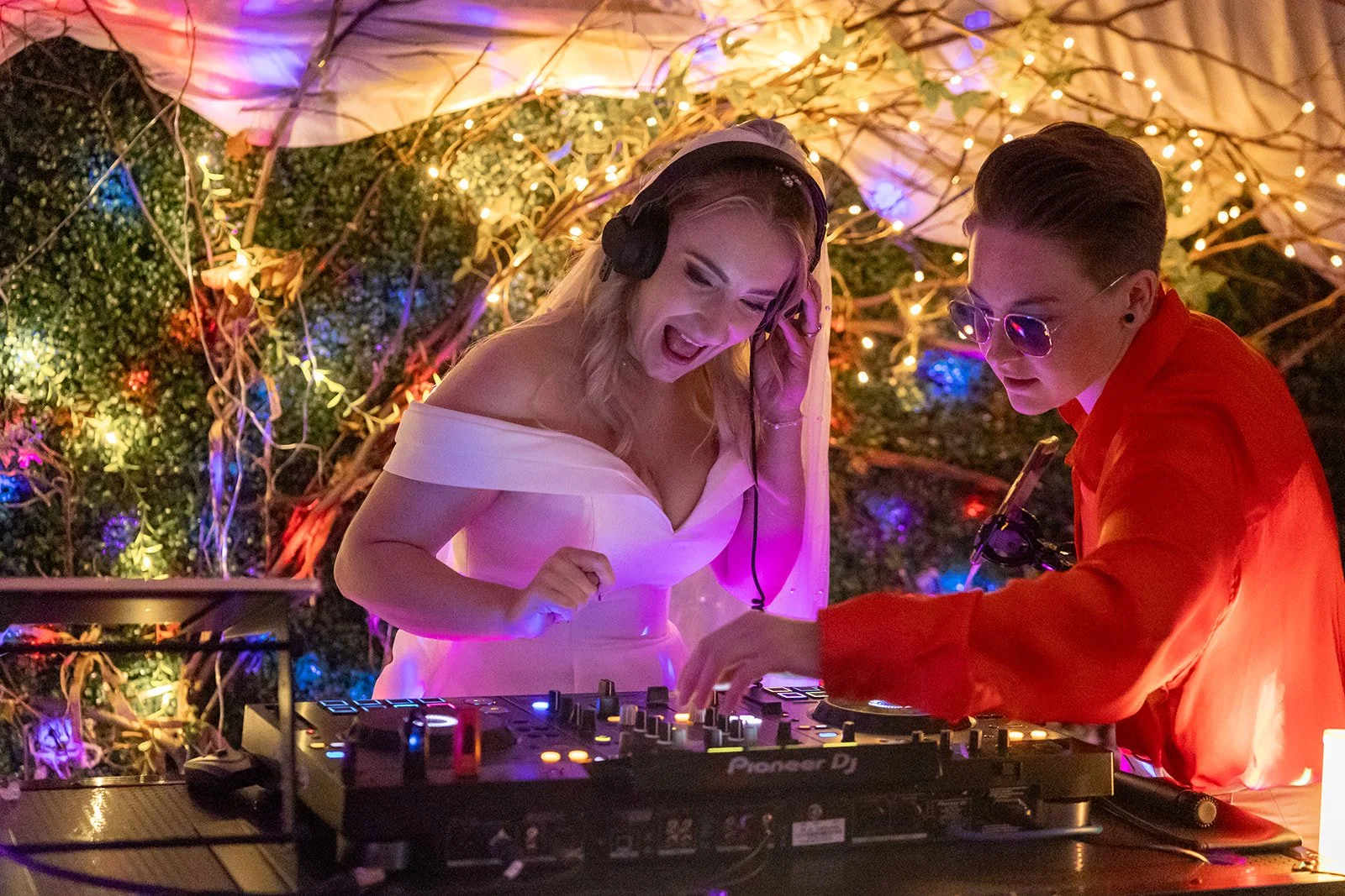 Two women behind the DJ booth. One woman in a white off-shoulder wedding dress and a DJ woman in an orange silk shirt and sunglasses. Both are concentrating and surrounded by colorful fairy lights and foliage, with a lively, energetic atmosphere