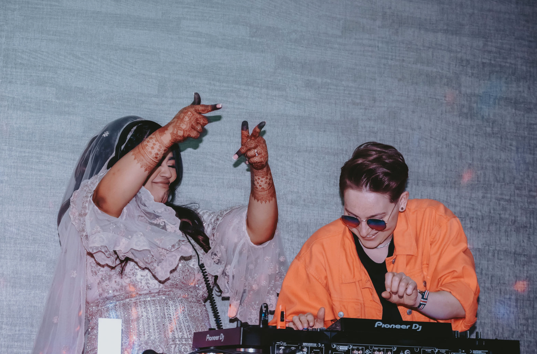 Two women dancing and listening to music with DJ equipment. One woman, the bride, wearing a jewelry, veil and henna. The other is the DJ, wearing sunglasses and an orange jacket denim jacket