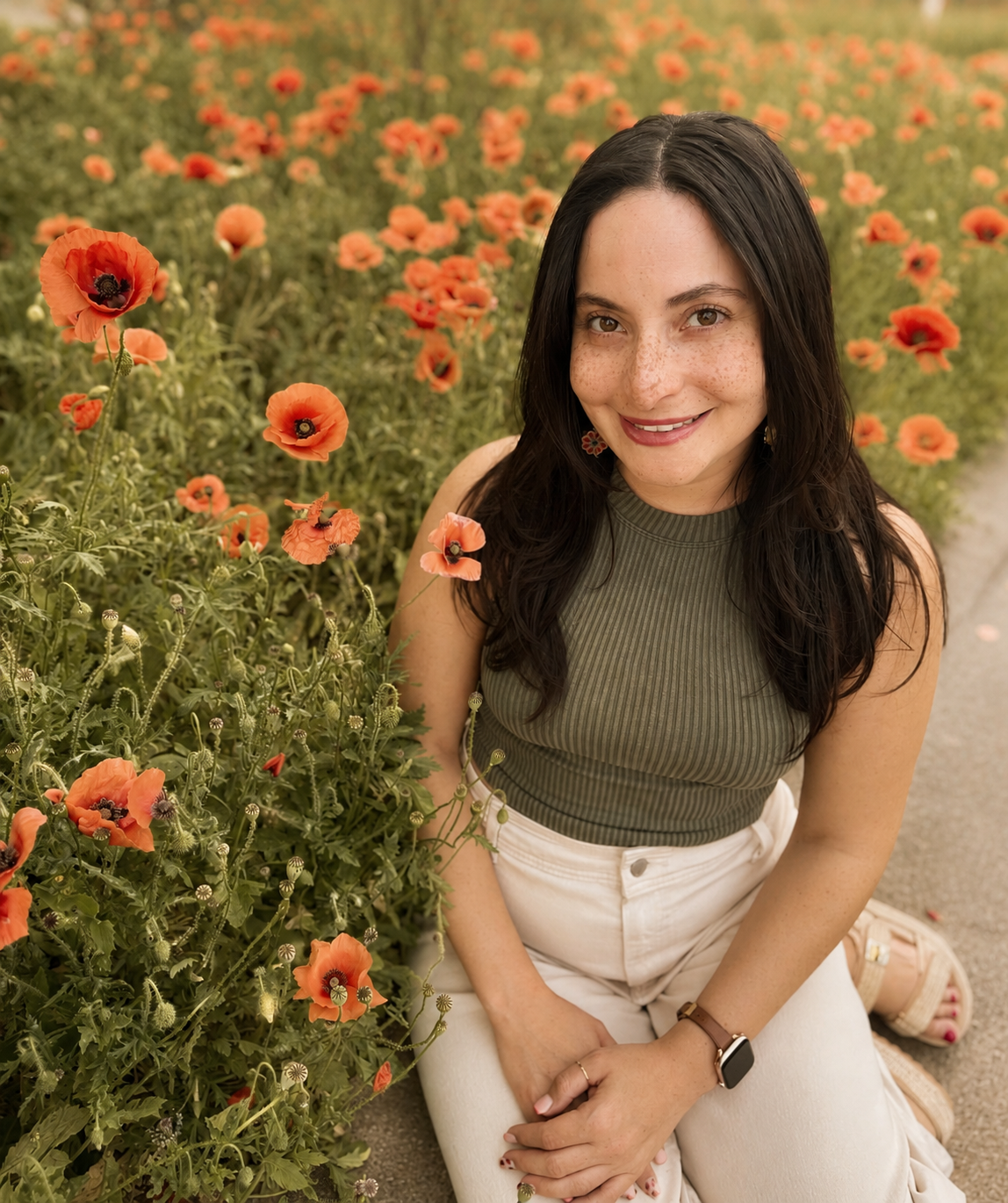 A woman with long dark hair and freckles, smiling, kneeling next to a field of orange poppies, wearing a green sleeveless top, white pants, and a smartwatch.