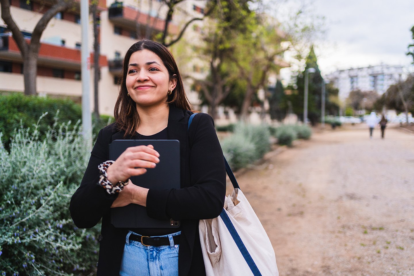 A young woman with dark hair, wearing a black blazer and light blue jeans, is standing outdoors in a park or walkway with trees, bushes, and buildings in the background. She is smiling and holding a closed laptop or tablet close to her chest.