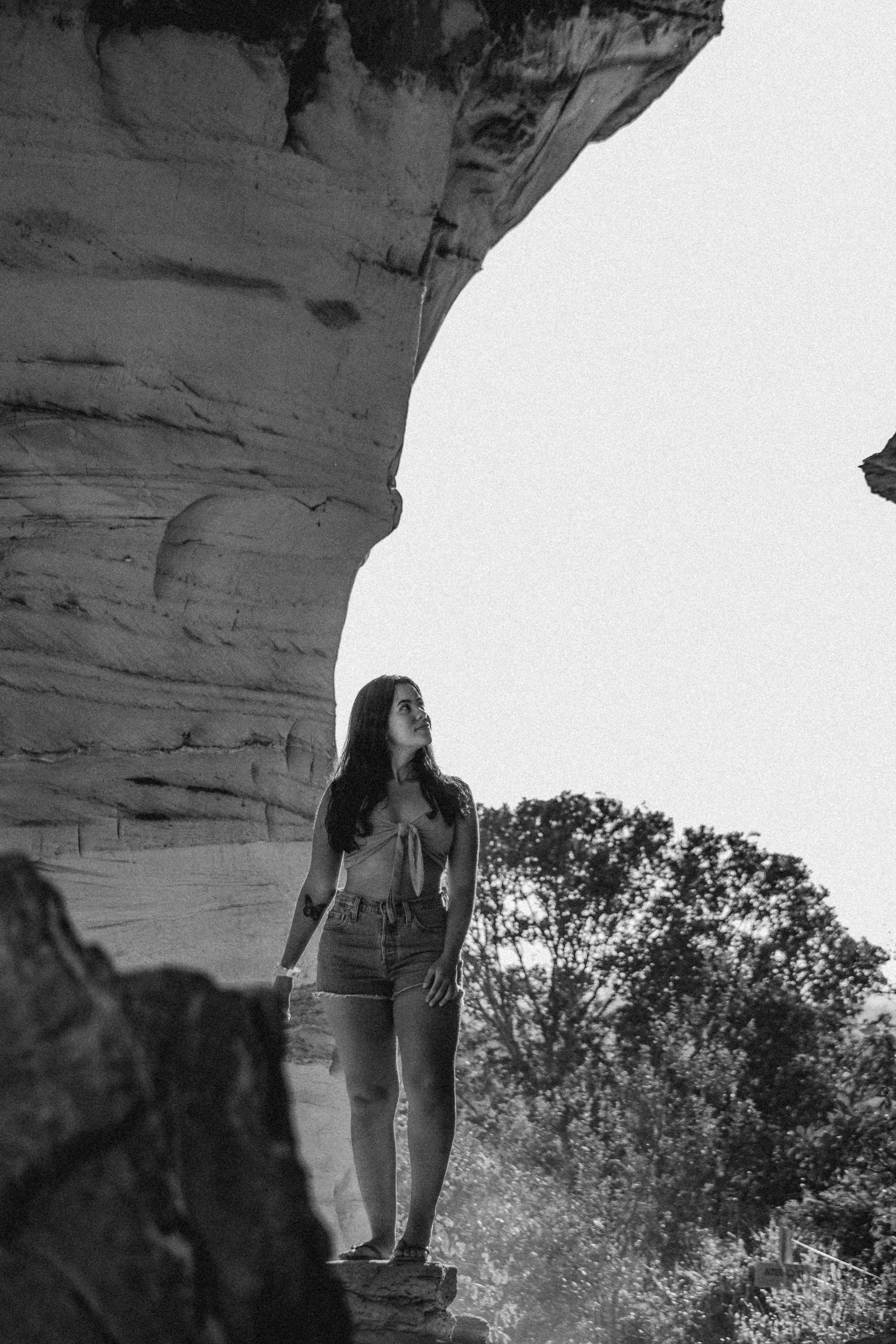 Jovem mulher de shorts jeans e top, de cabelos longos, em pé ao ar livre, olhando para cima, com uma formação rochosa ao fundo e árvores ao fundo, em uma cena em preto e branco.