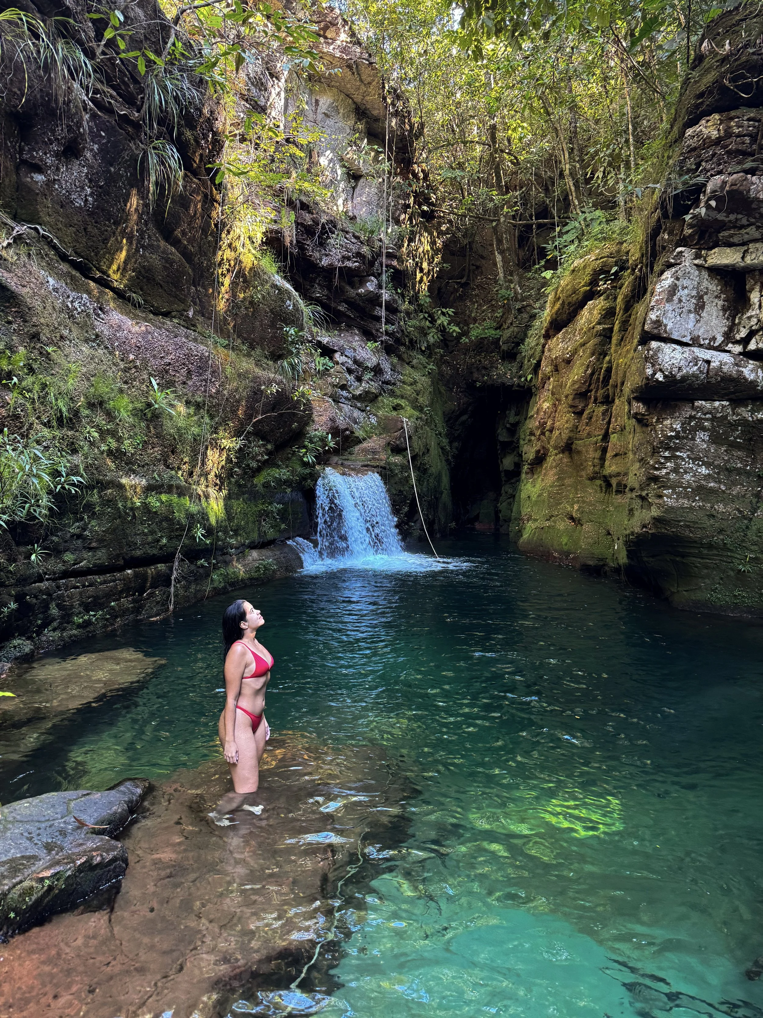 Mulher de biquíni vermelho de pé na água de uma cachoeira em uma floresta densa e verdejante, com rochas e o fluxo de água ao fundo.