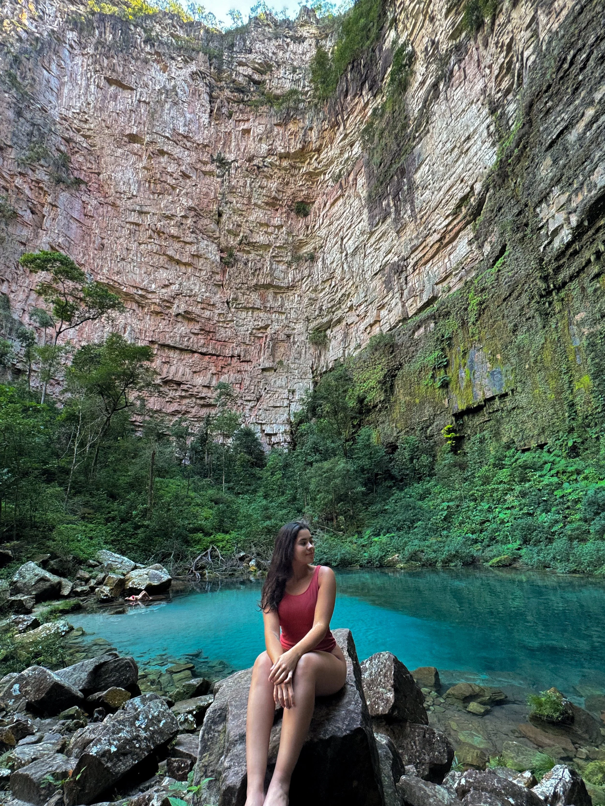 Mulher de vestido vermelho sentado em uma pedra ao lado de uma água azulada em um cânion ou ravina com paredes de rocha e vegetação ao redor.