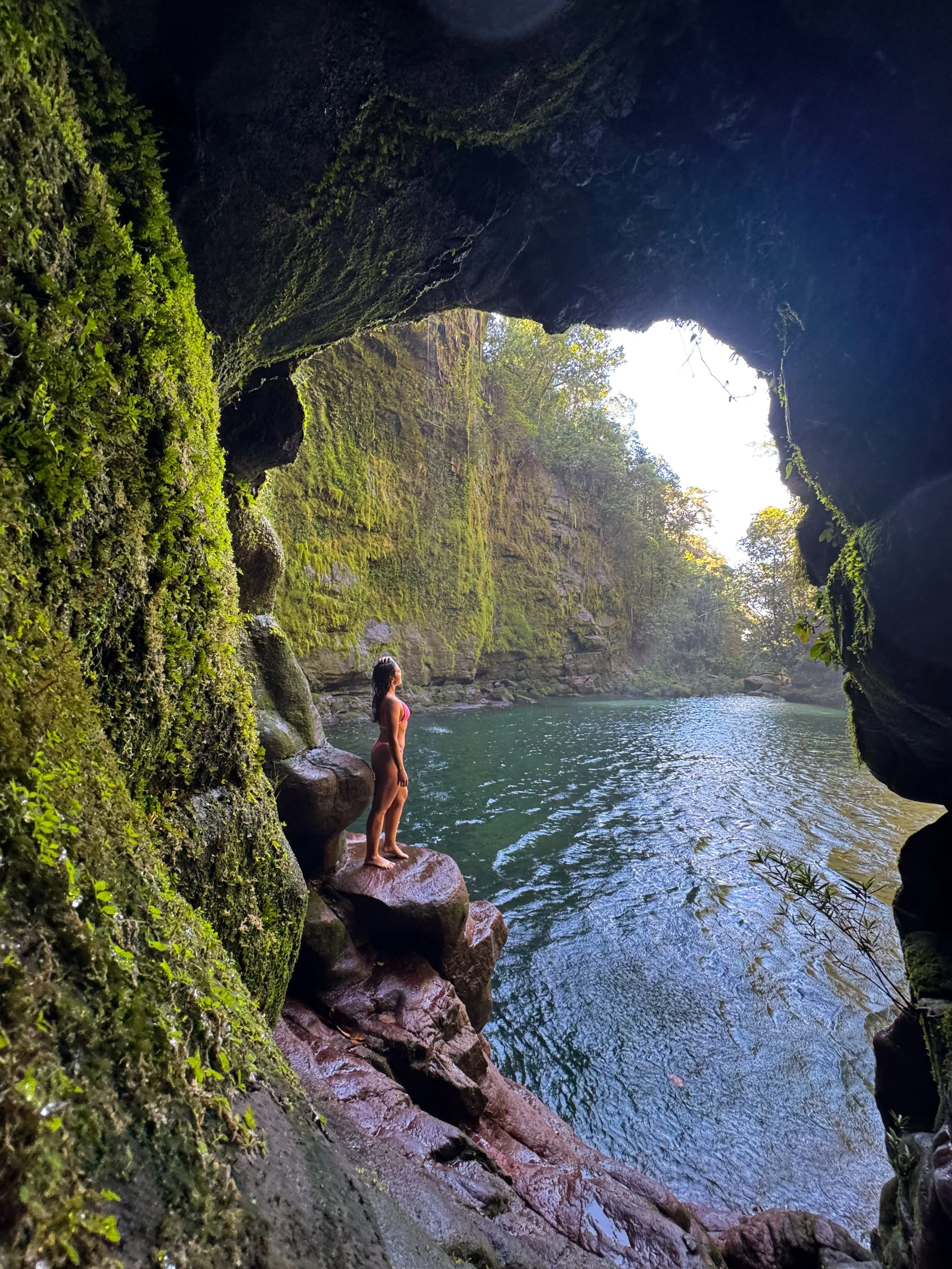 Mulher de biquíni de pé em uma rocha dentro de uma caverna ao lado de um rio natural rodeado por vegetação verdejante.