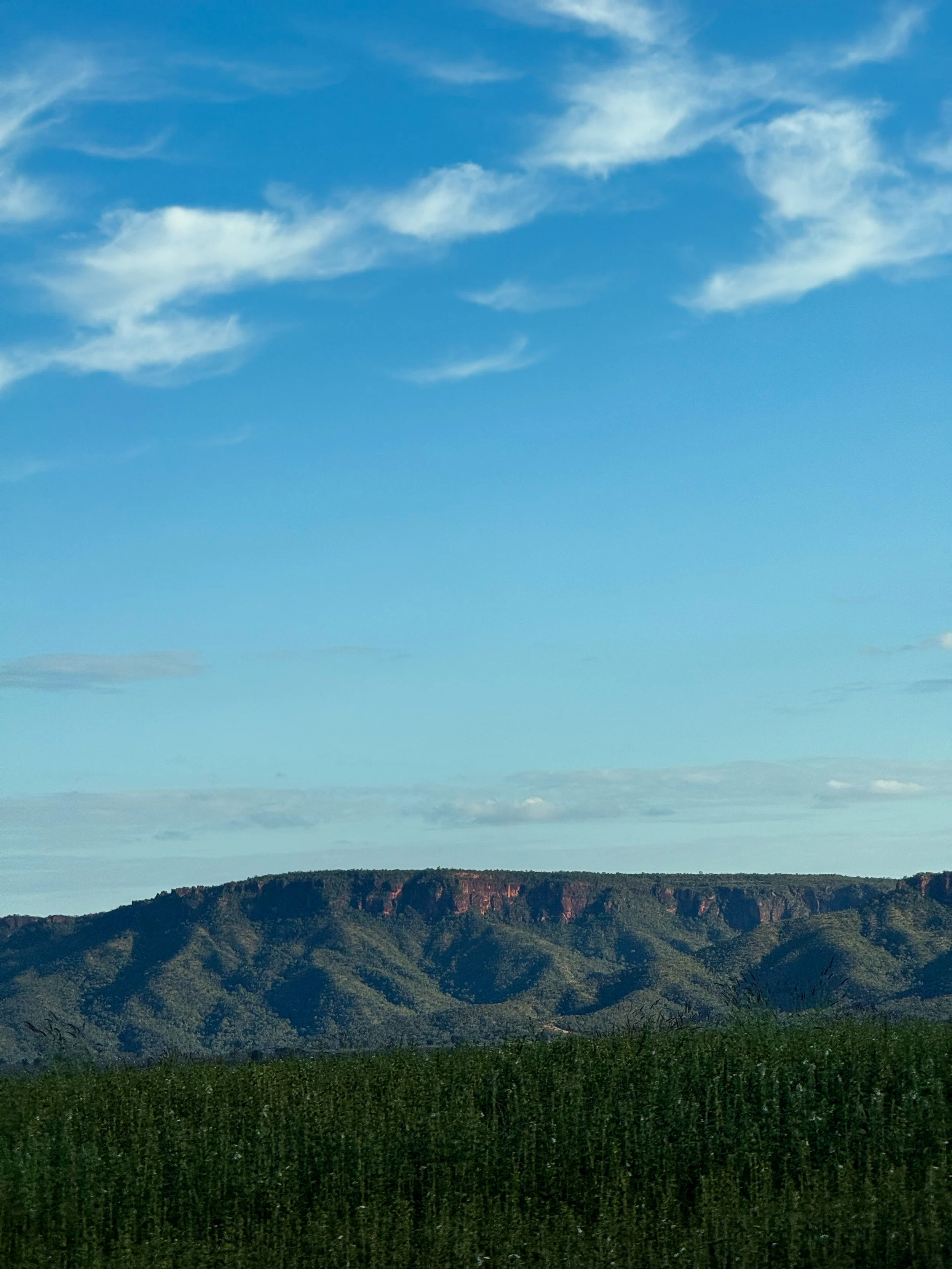 Vista de uma cadeia de montanhas verdes sob um céu azul com algumas nuvens brancas.