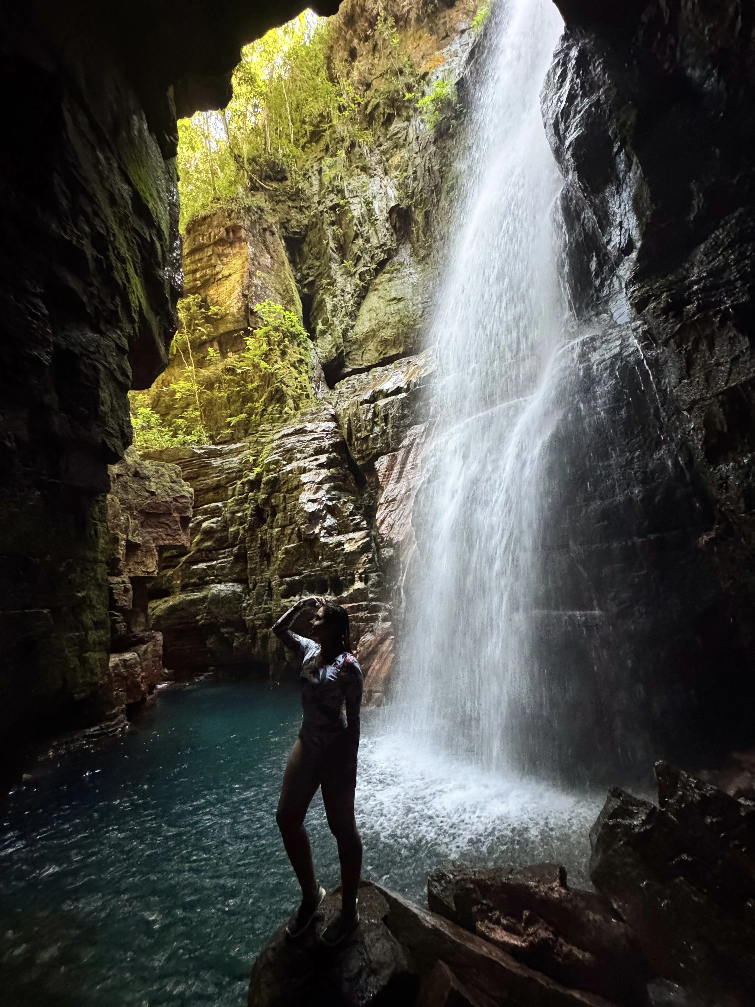 Pessoa de standing em uma rocha dentro de uma caverna, observando uma cachoeira e a água caindo de uma rocha alta, com vegetação ao redor.