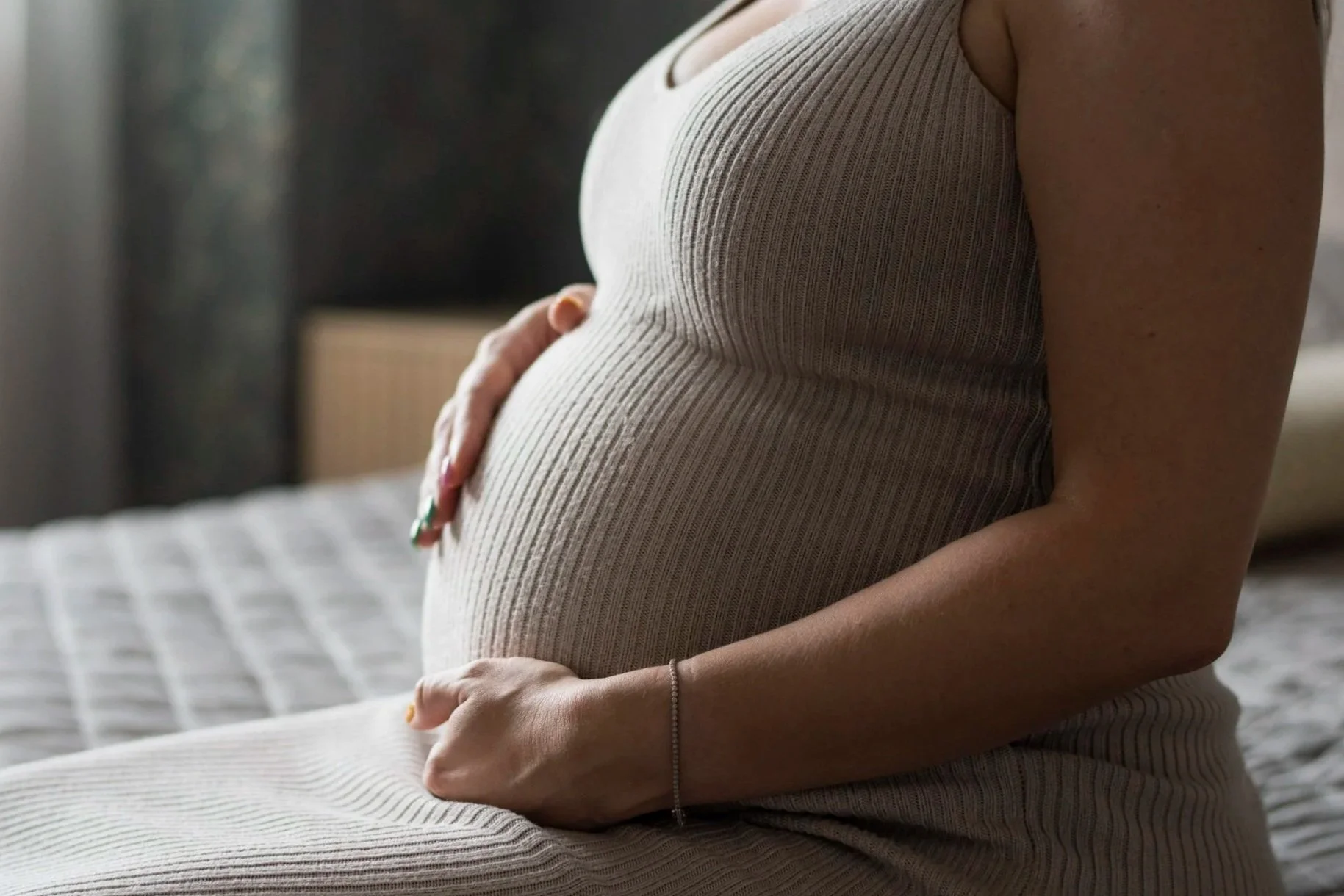 A pregnant woman sitting on a bed with her hands gently resting on her belly. She is wearing a sleeveless, ribbed beige top. The background shows a bed with a #checked sheet and some blurred furniture.