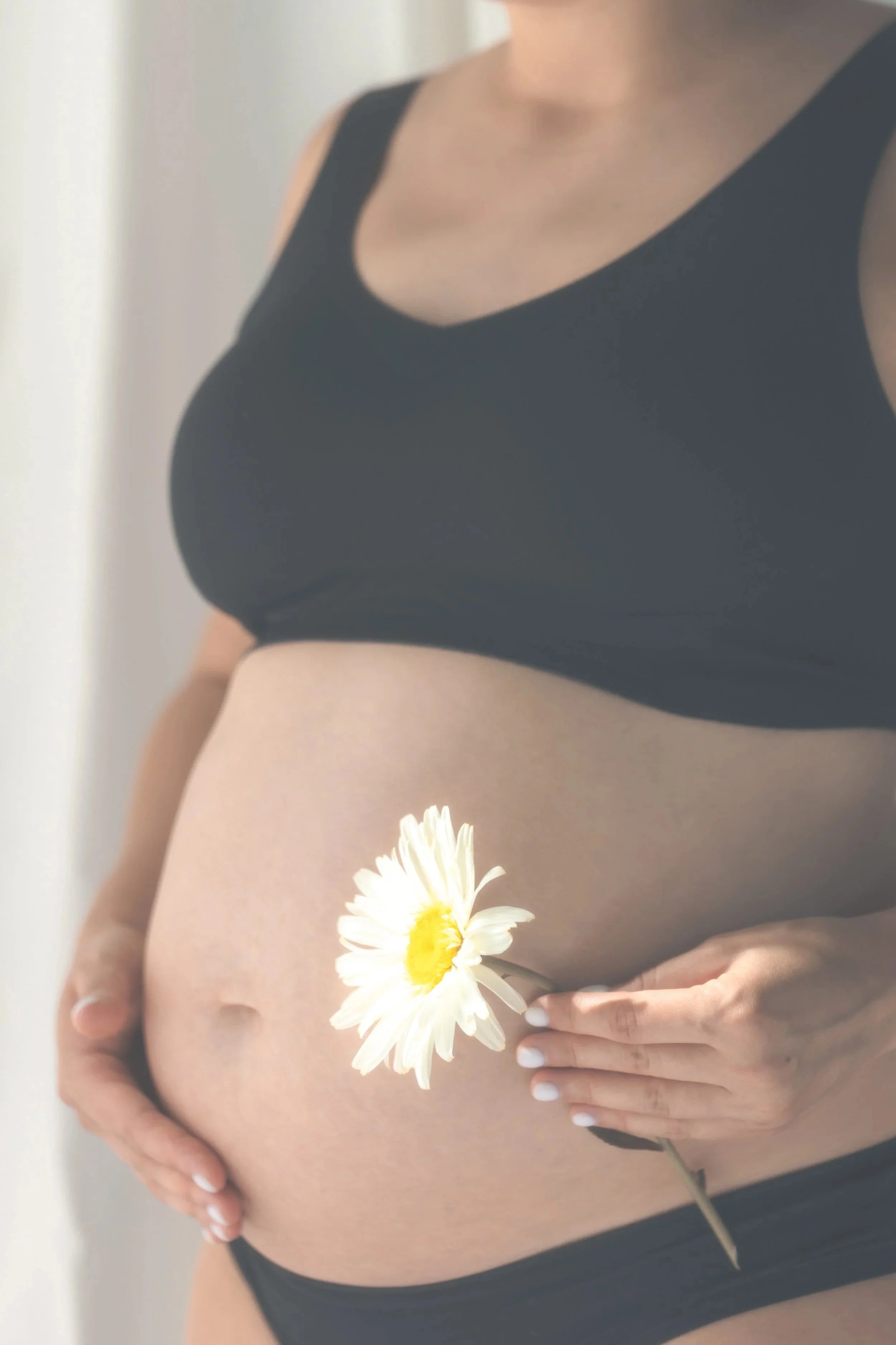 A pregnant woman wearing a black bra, holding a white flower with a yellow center near her lower belly.