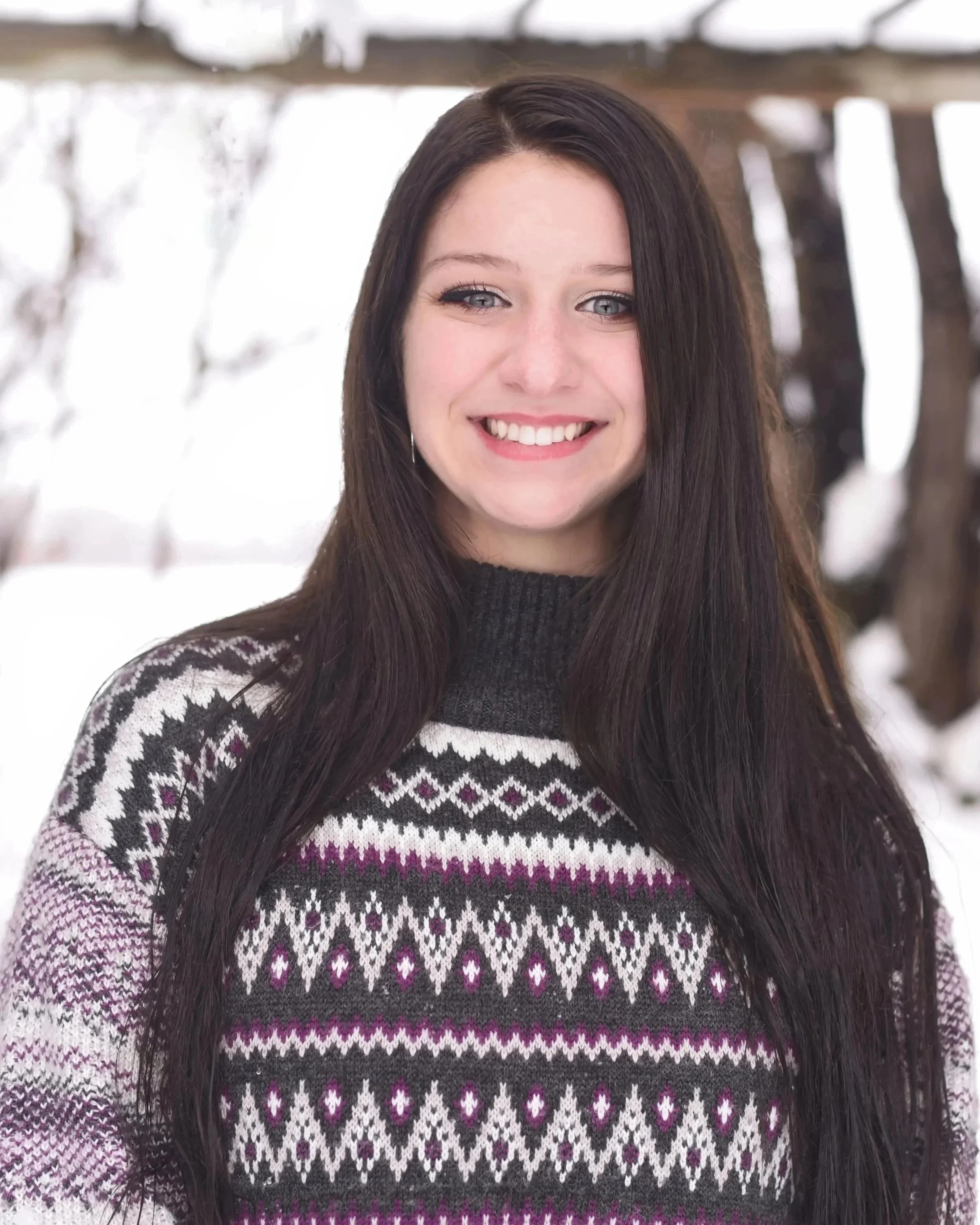 A smiling woman with long dark hair, wearing a patterned sweater, standing outdoors in a winter setting with snow and trees in the background.