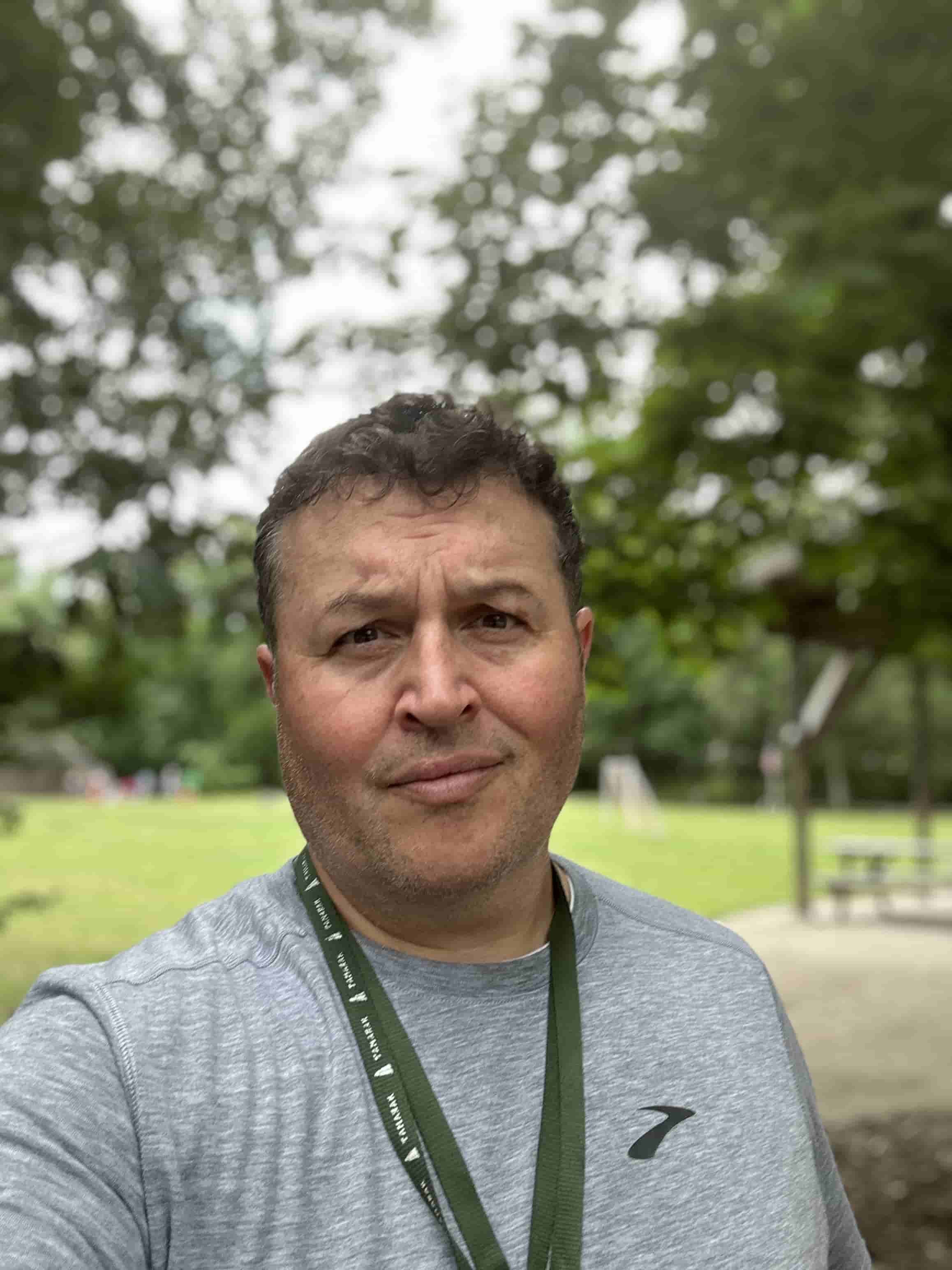 A man with dark curly hair and a gray athletic shirt taking a selfie outdoors with a grassy park, trees, and a picnic table in the background.