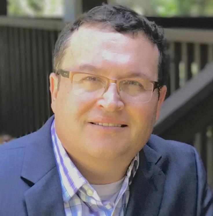A man with glasses and short dark hair smiling outdoors, wearing a suit jacket and a checkered shirt.