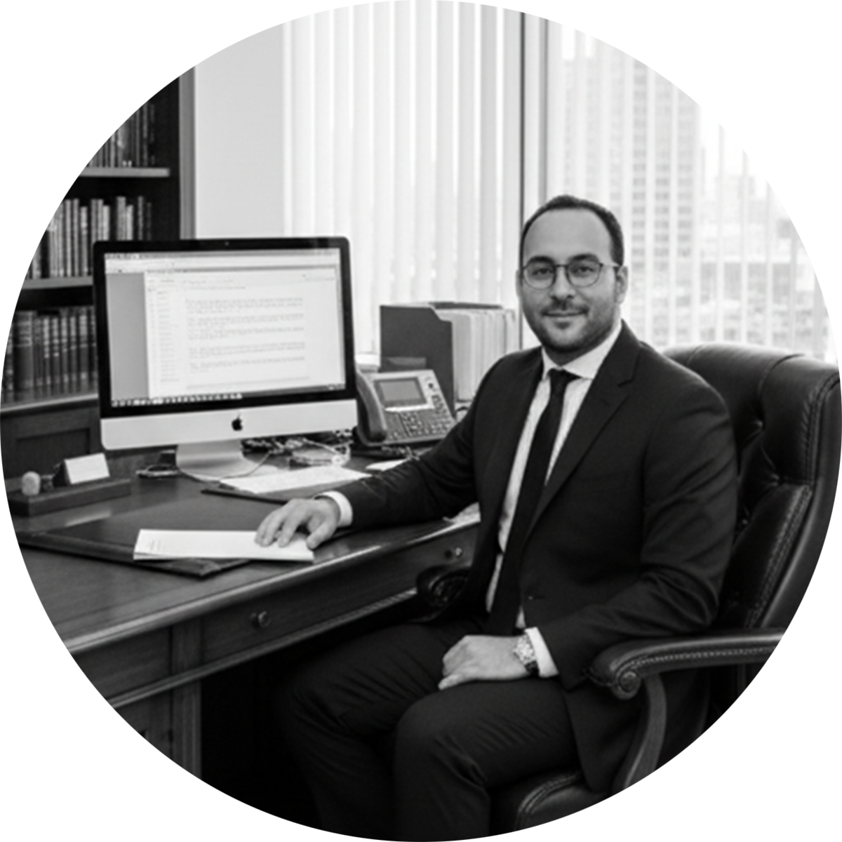 A man in a business suit sitting at a desk in an office, with a desktop computer, phone, and office supplies, looking at the camera.