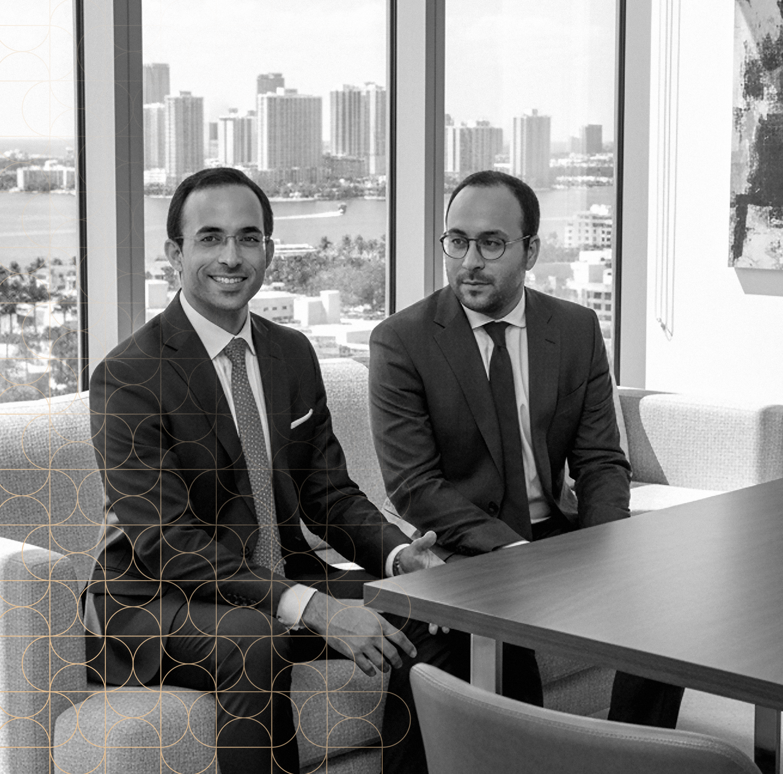Two men in suits sitting at a conference table in an office with large windows overlooking a city skyline.