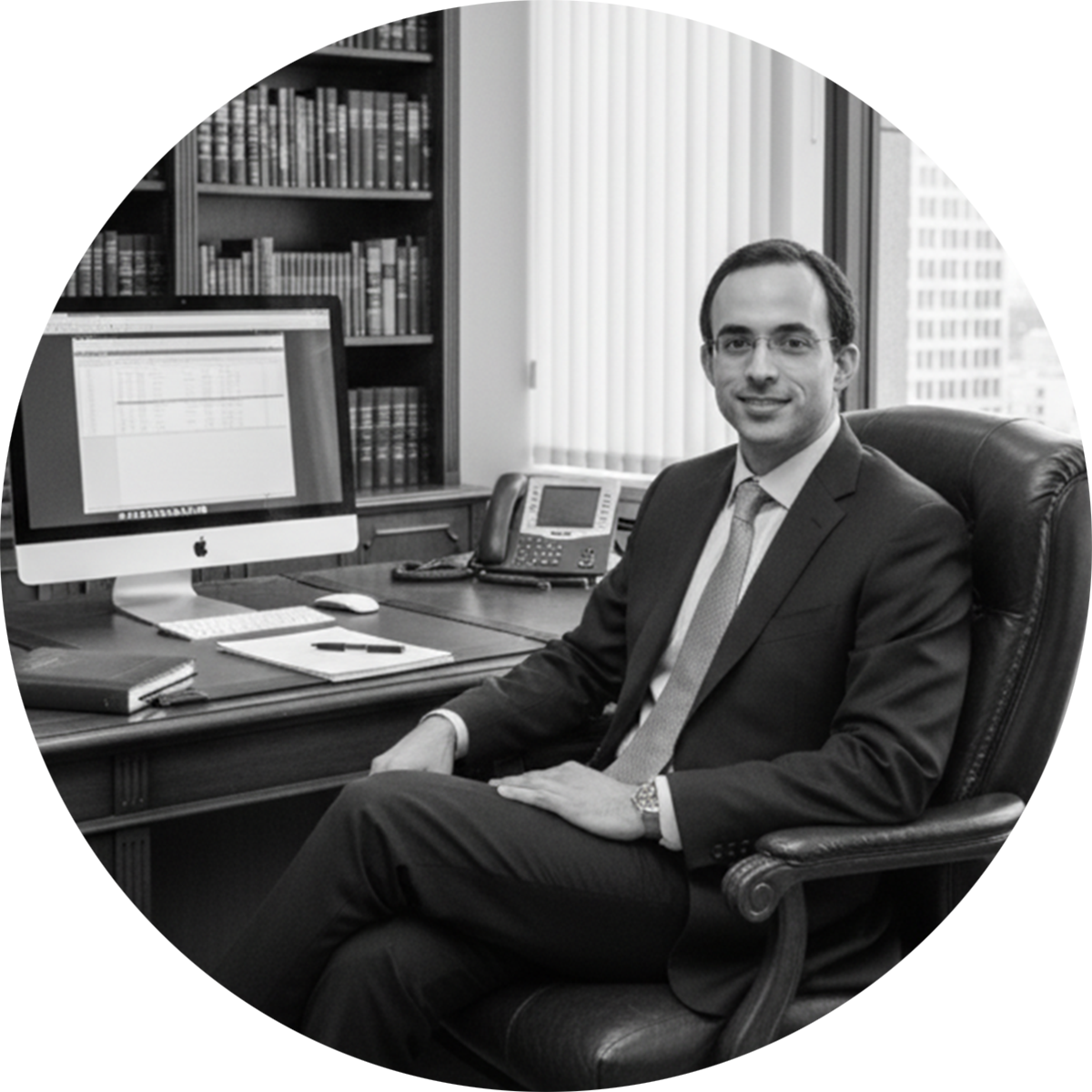 A man wearing glasses in a suit sitting in an office chair at a desk with a computer, bookshelves, and office phone visible behind him.
