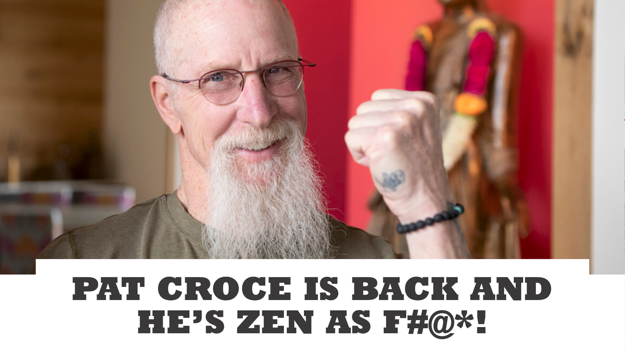 A smiling Pat Croce with a white beard and glasses making a fist pump gesture. The background features a colorful wooden sculpture and red wall.