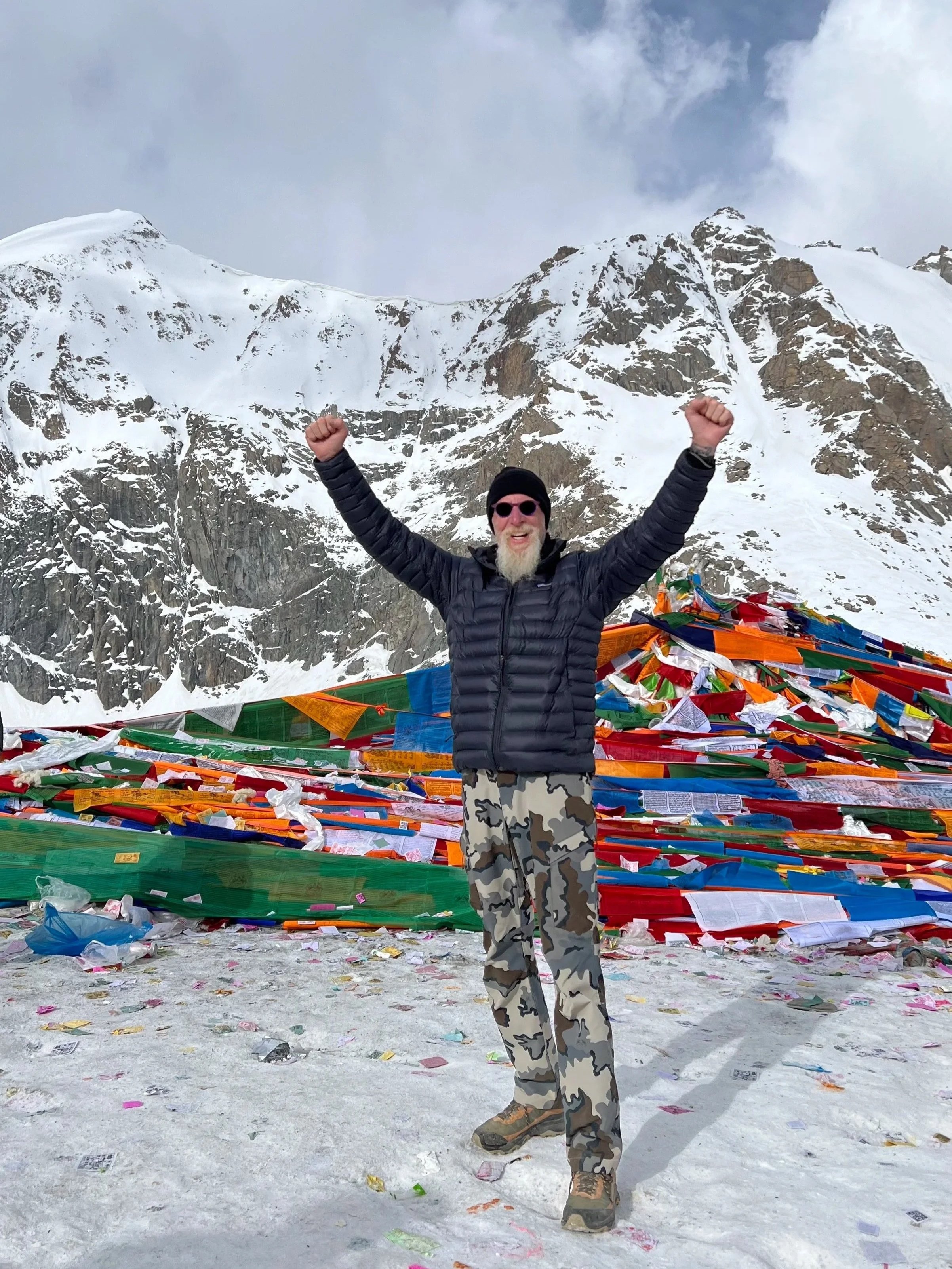 Photograph of Pat Croce with a white beard and sunglasses, wearing a black jacket, camouflage pants, and a black beanie, celebrating on Mount Kailash in Tibet, with colorful prayer flags in the background..