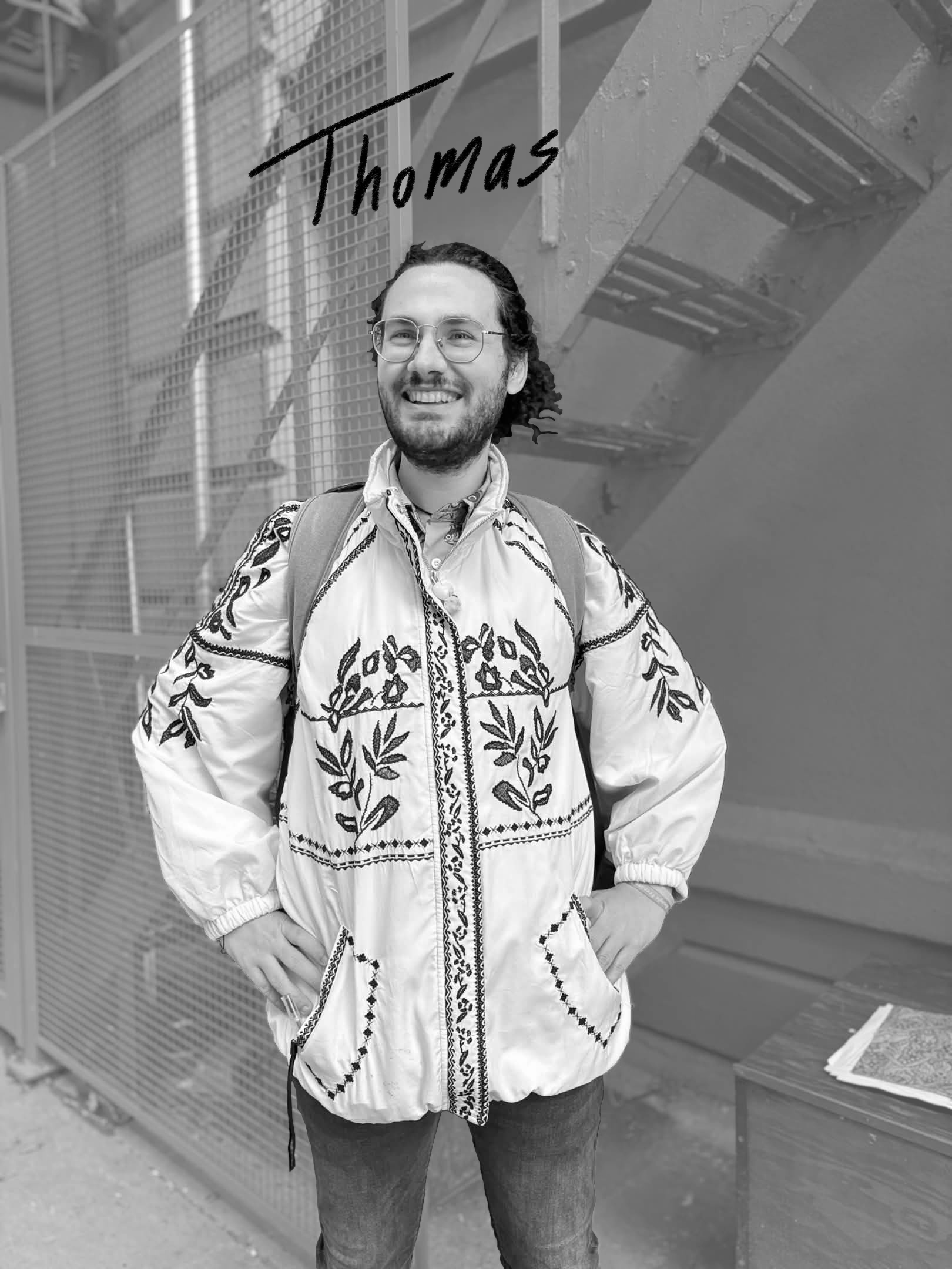 A smiling man with glasses and curly hair standing outdoors, wearing a patterned jacket and a backpack, standing in front of a metal fence with an outdoor staircase behind him, smiling with his hands on his hips.