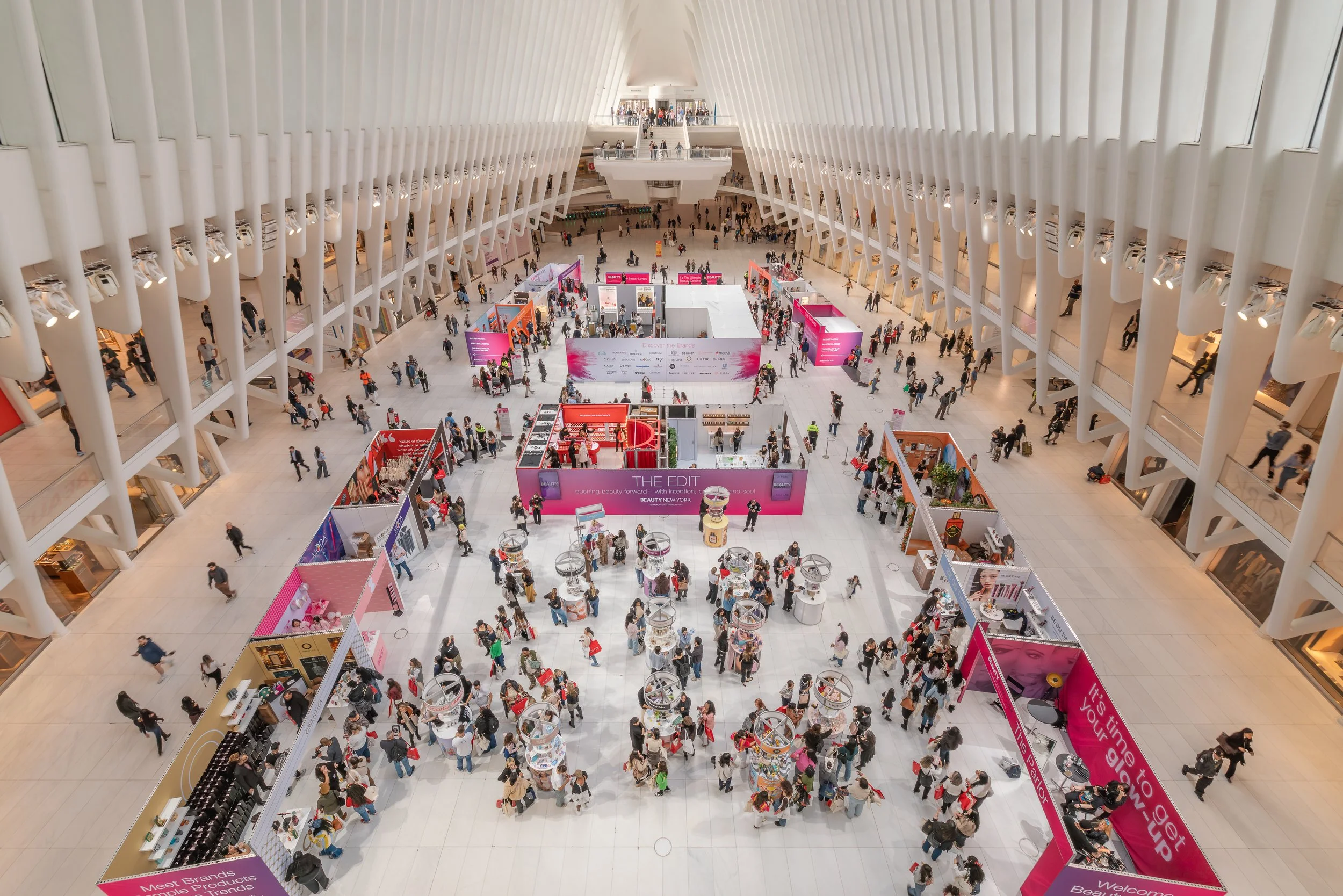A large indoor space with high ceilings and curved white walls, filled with numerous booths and many visitors walking around, suggesting a trade show or exhibition event.