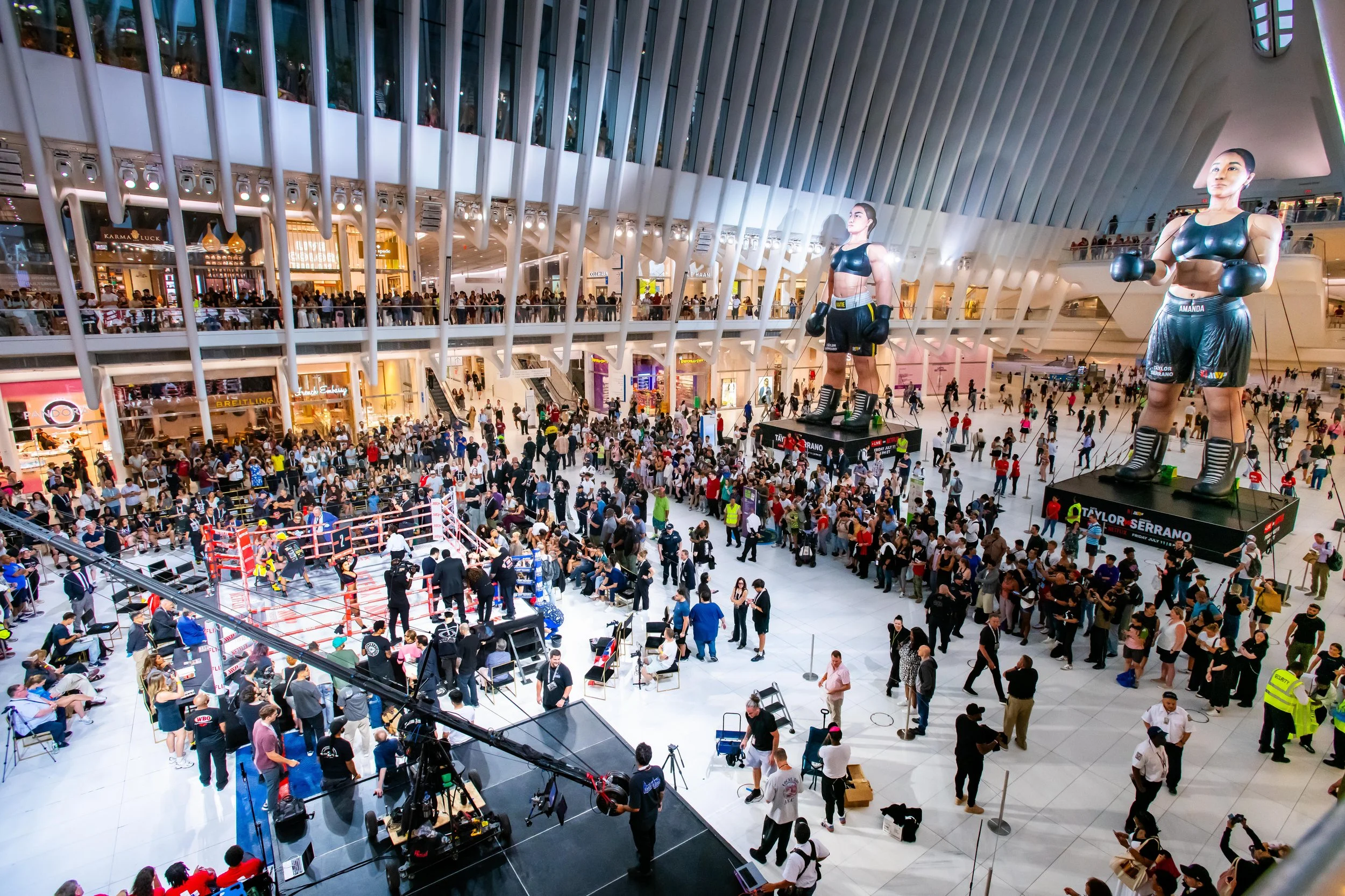 People gathered in a mall for a boxing event with a ring and oversized boxing statue displays.