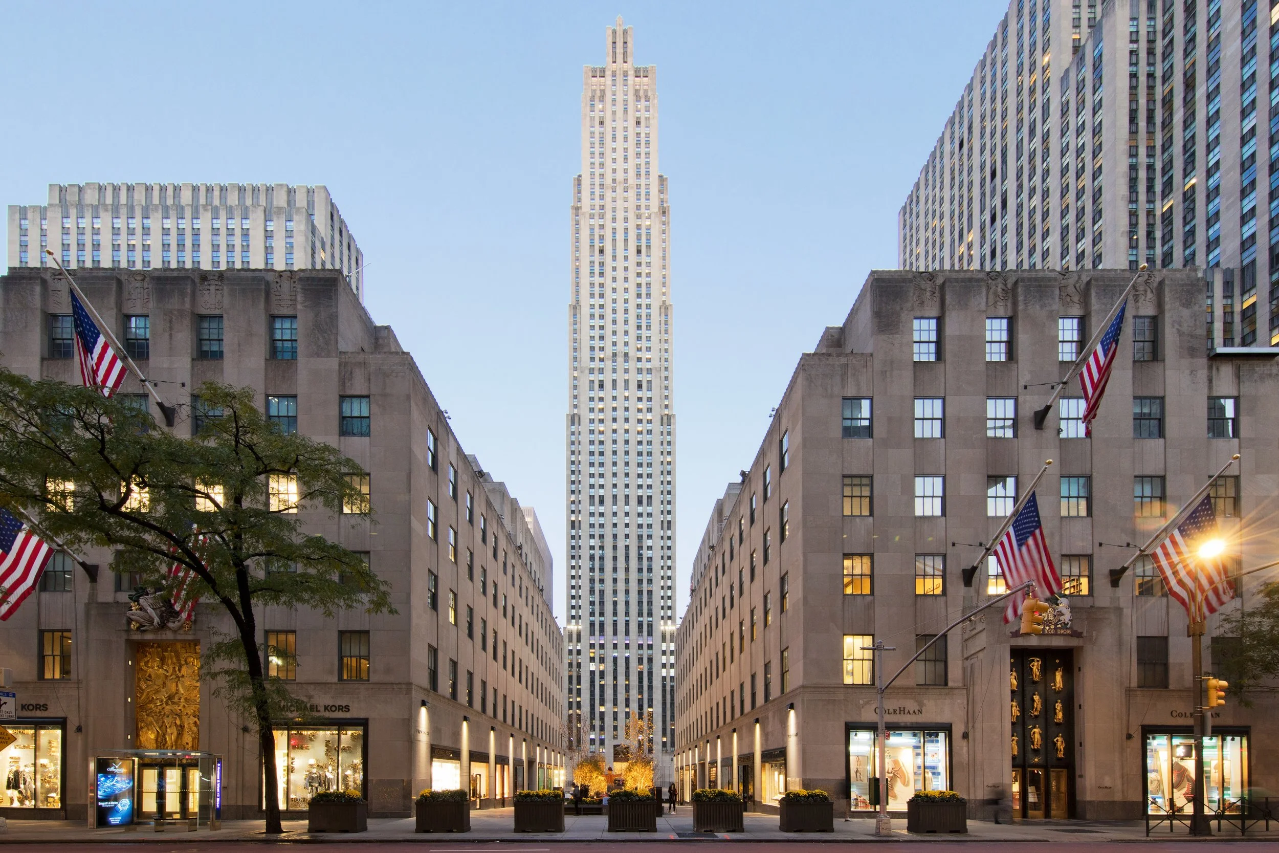 Tall skyscraper between two shorter buildings with retail stores at street level, American flags on poles, streetlights, trees, and a clear blue sky.