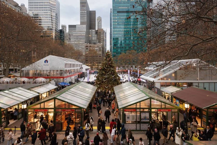 A bustling holiday market in a city square with a decorated Christmas tree at the center, surrounded by glass-roofed booths, tall buildings, and leafless trees.