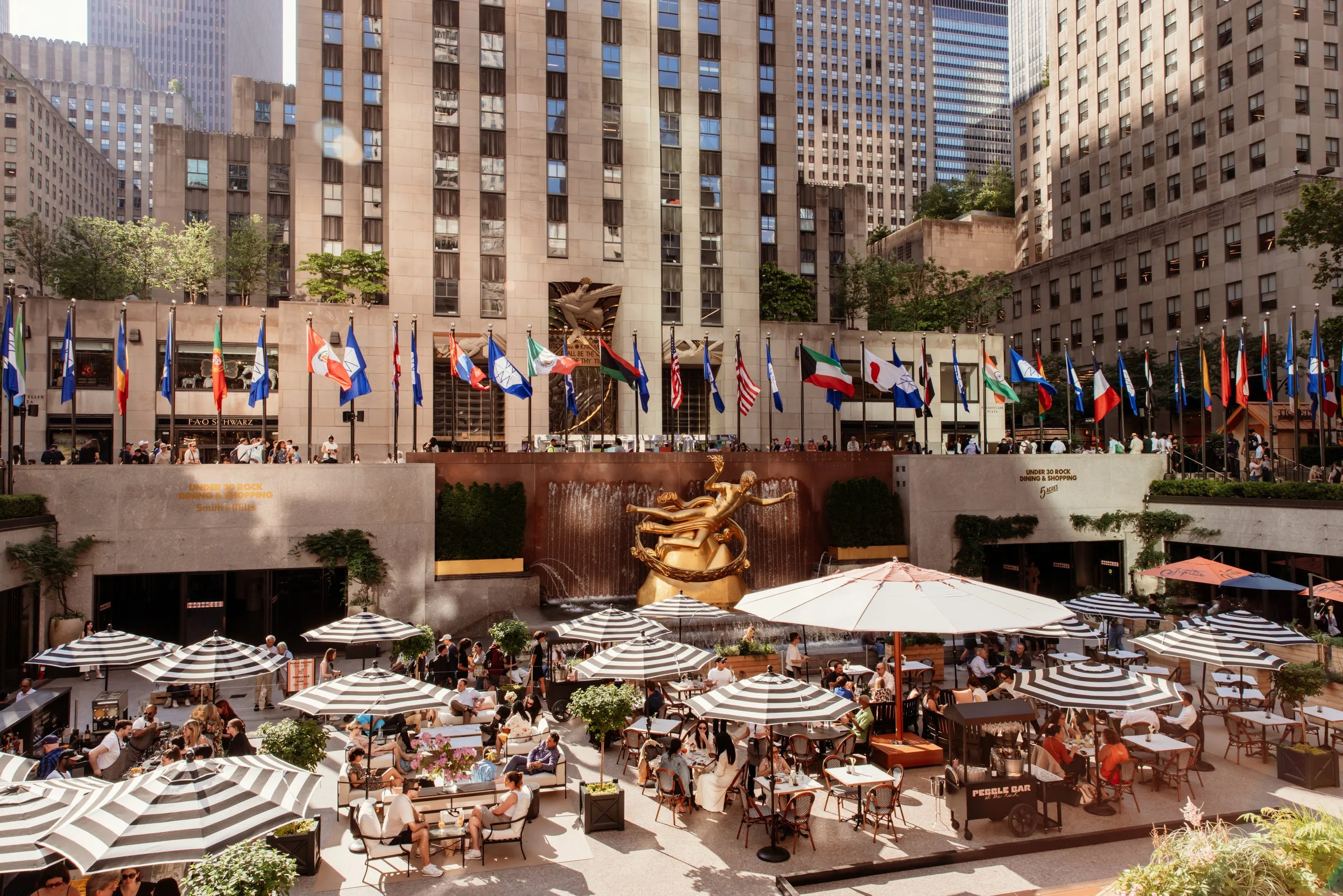 An outdoor plaza in a city with numerous people sitting at tables under umbrellas. Behind them is a large fountain with a golden sculpture of a woman and water flowing, surrounded by flags of various countries. Tall buildings serve as a backdrop.