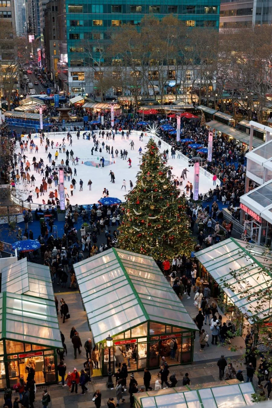 People ice skating on an outdoor rink decorated with Christmas trees in a busy urban area during the evening. Market stalls and bright city lights surround the scene.