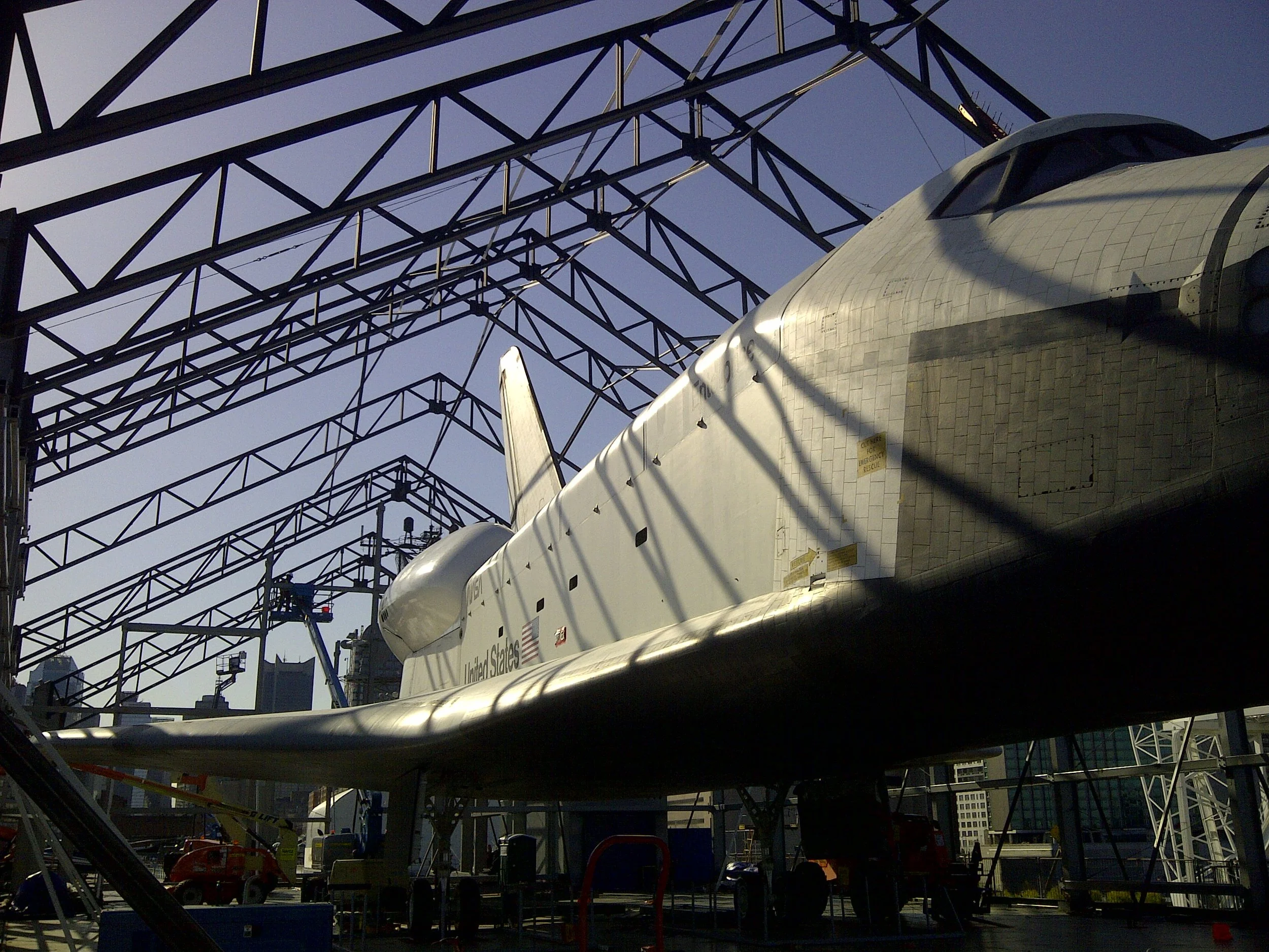 An aircraft, possibly a space shuttle, is inside a large hangar or structure made of metal beams. The aircraft's body is metallic and reflective, with a shadow cast on it. In the background, there are cranes, construction equipment, and a cityscape with tall buildings.