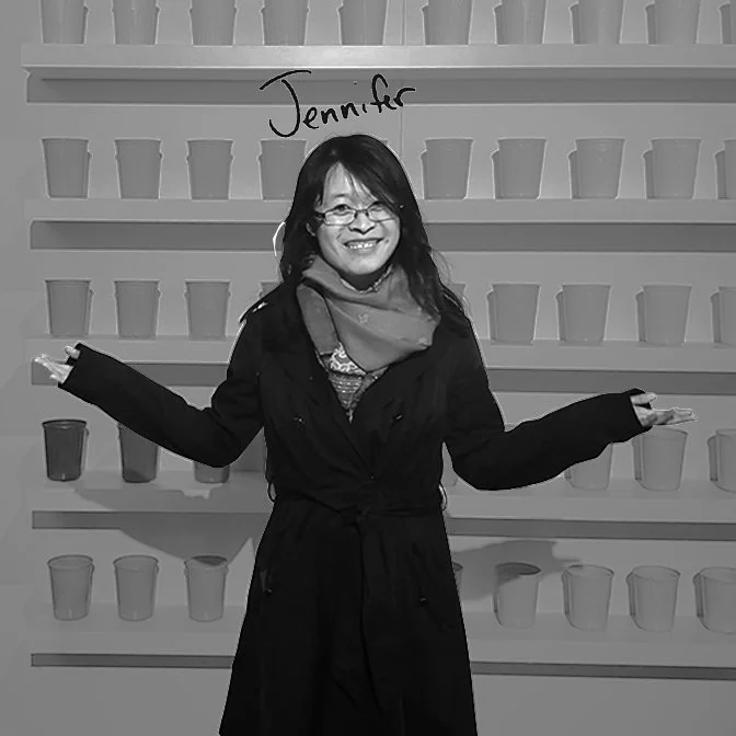 Woman with glasses and scarf standing in front of a wall with multiple plant pots, smiling with arms outstretched.