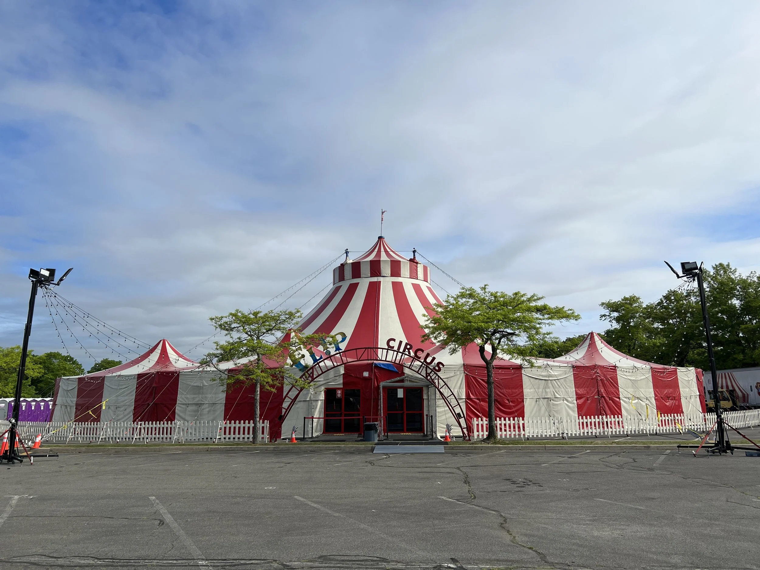 A red and white striped circus tent set up in an empty parking lot with trees and floodlights around it.
