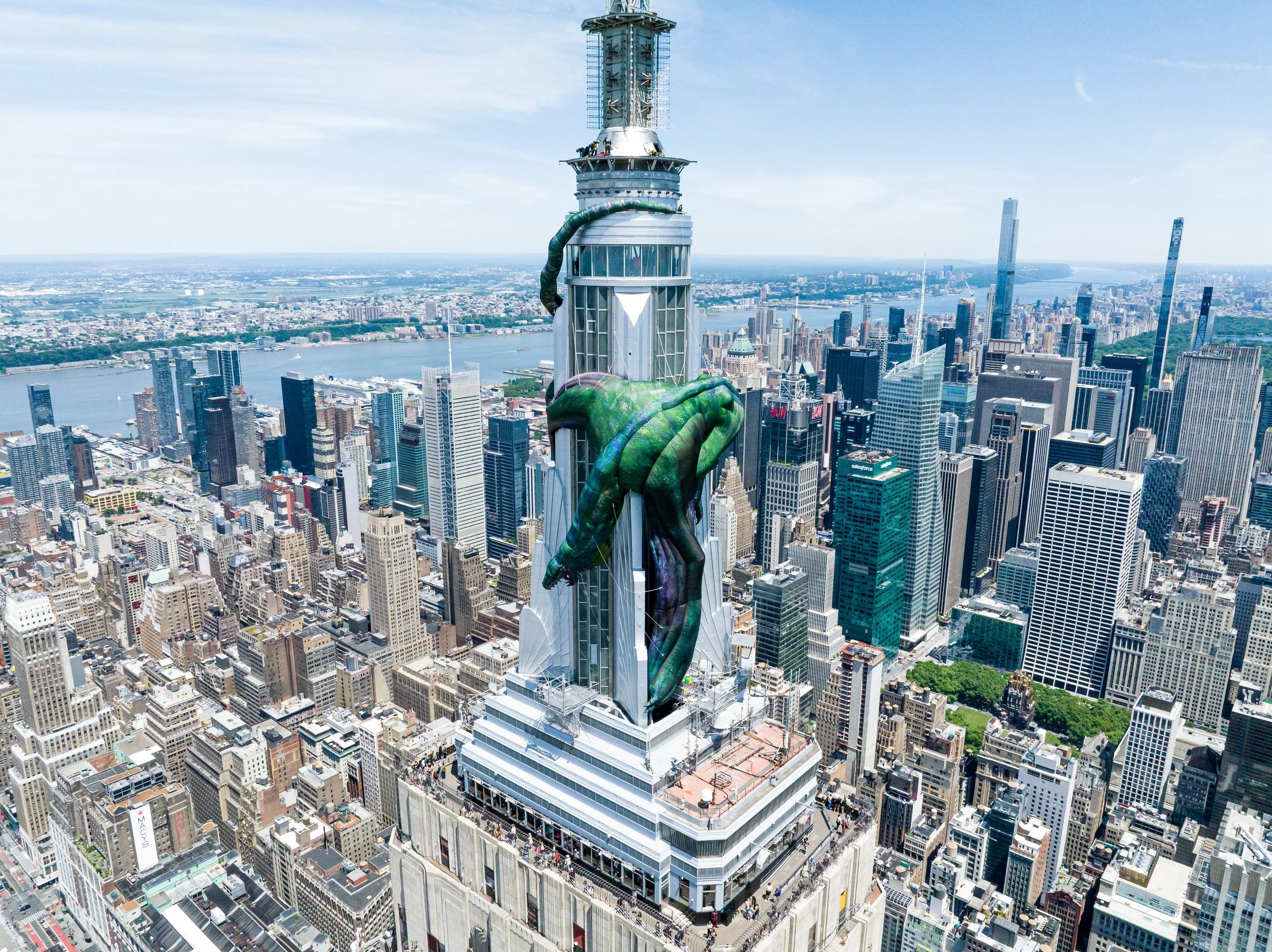 A tall skyscraper in New York City with a large green artistic sculpture of a hand wrapping around the building. The city skyline and river are visible in the background.