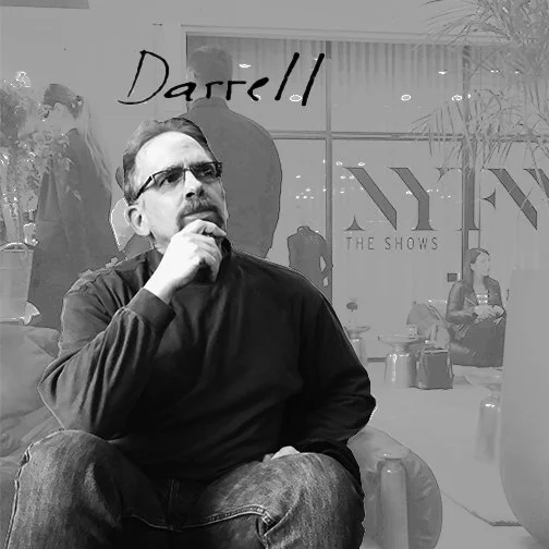 Man wearing glasses and black shirt sitting with his hand on his chin inside a shopping mall, with large glass windows and store displays in the background.