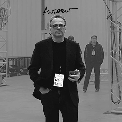 Man in glasses and black blazer holding a glass, standing in front of a camera mirror at an event.