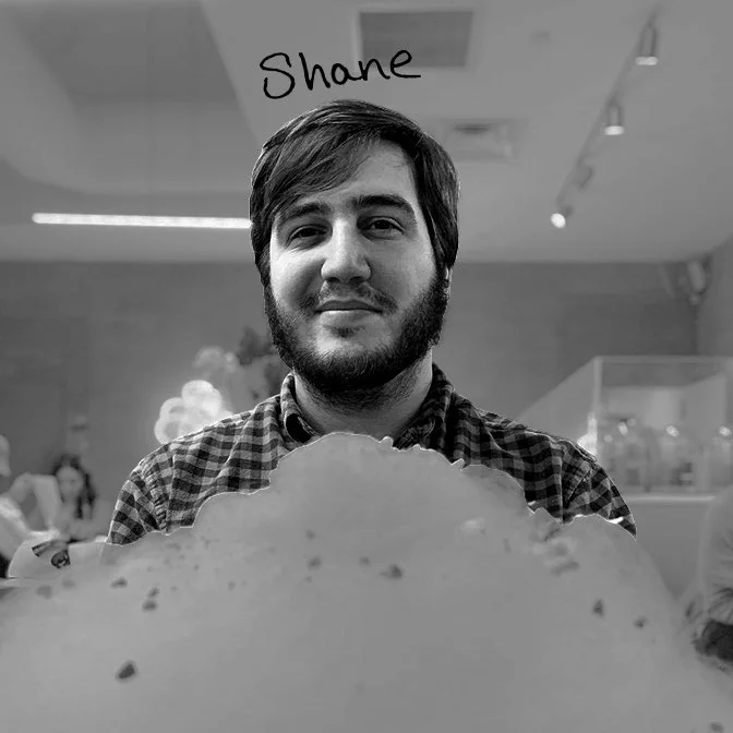 A young man with a beard and short hair smiling at the camera. The background suggests an indoor setting with other people.