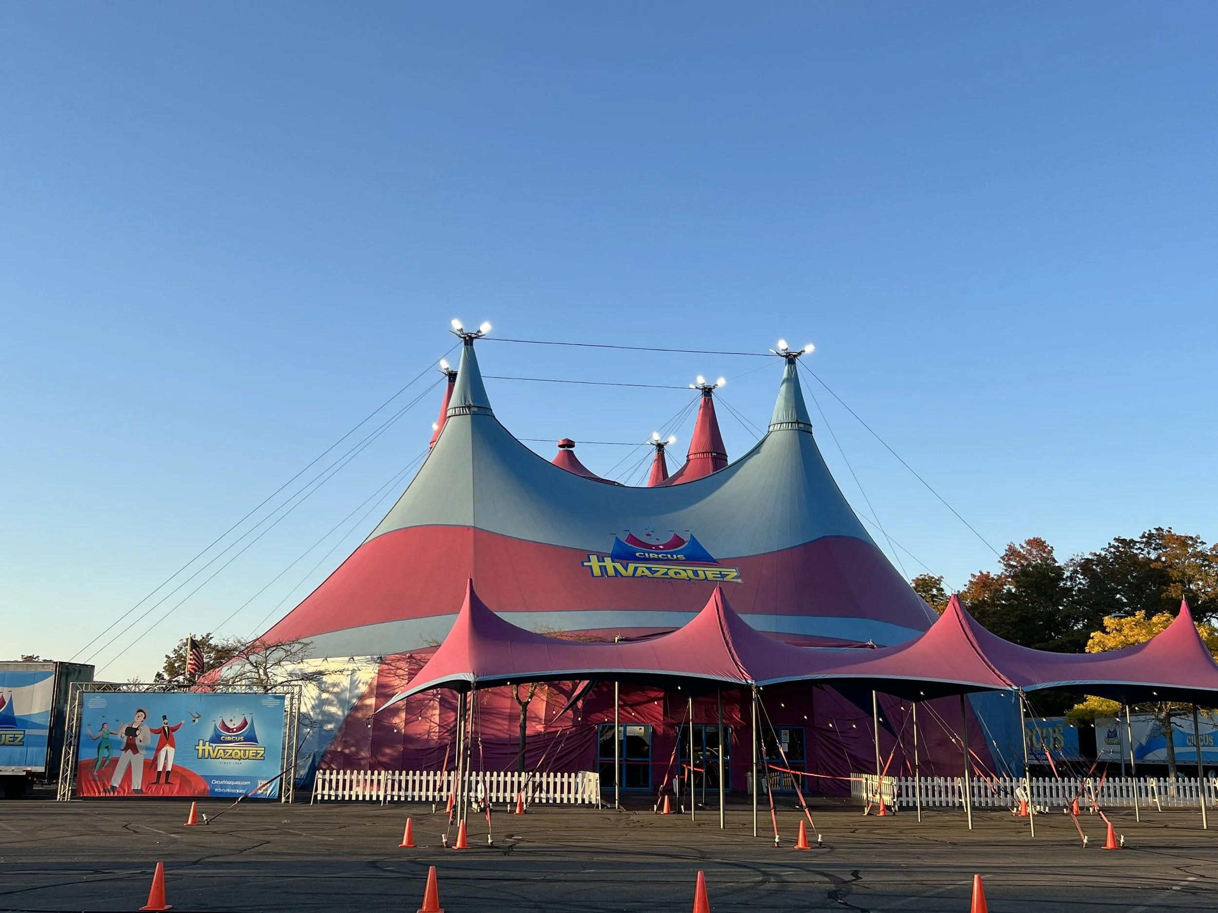 A colorful circus tent with pink, green, and red stripes and pointed peaks, set up in an outdoor area with safety cones and a white fence, under a clear blue sky.