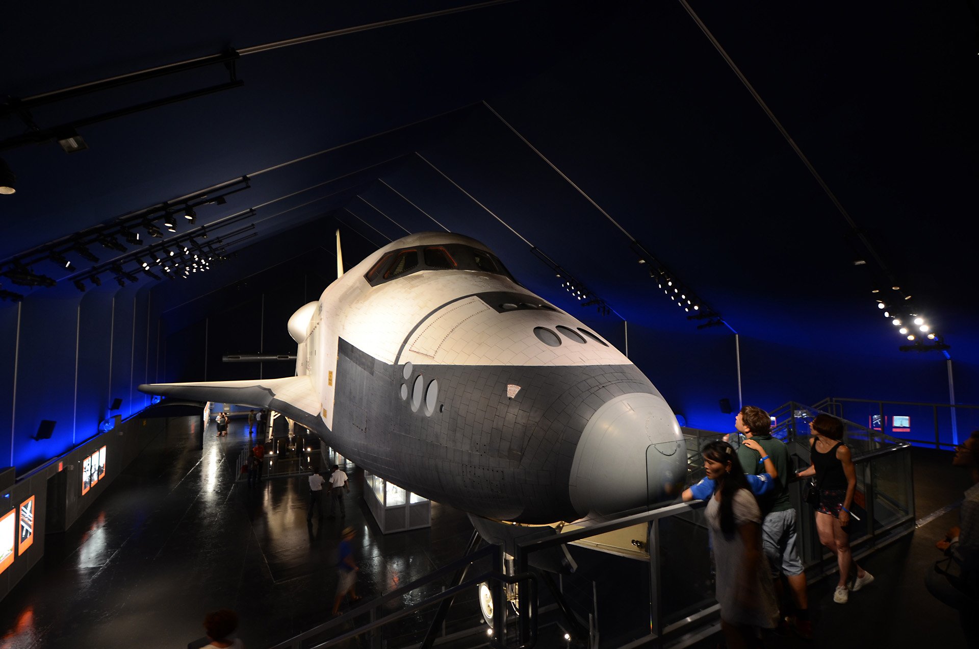 A space shuttle on display inside a museum with visitors examining it.