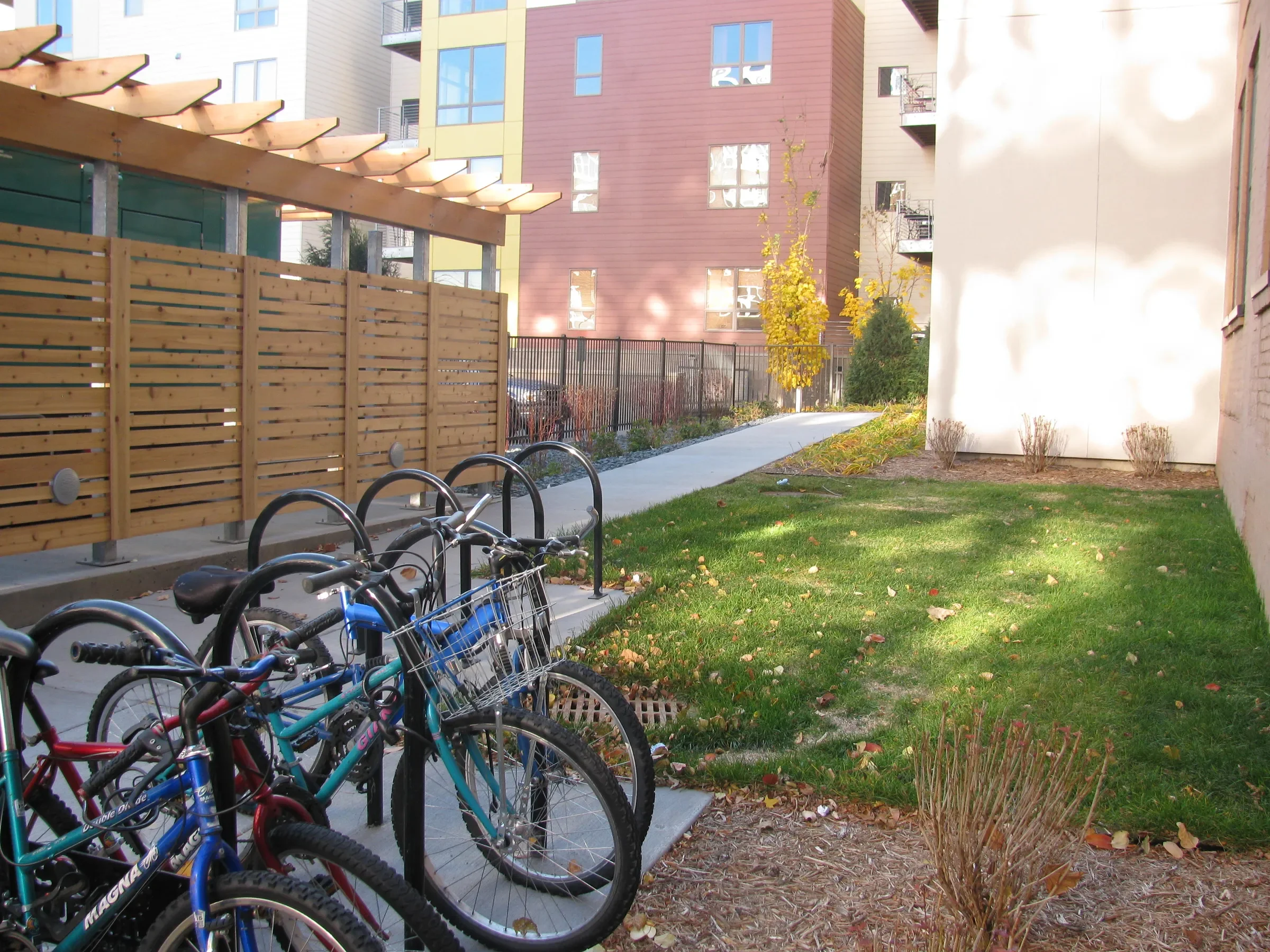 Bicycle parking area with bikes, wooden privacy fence, grassy yard, sidewalk, and multi-story modern apartment buildings in the background.