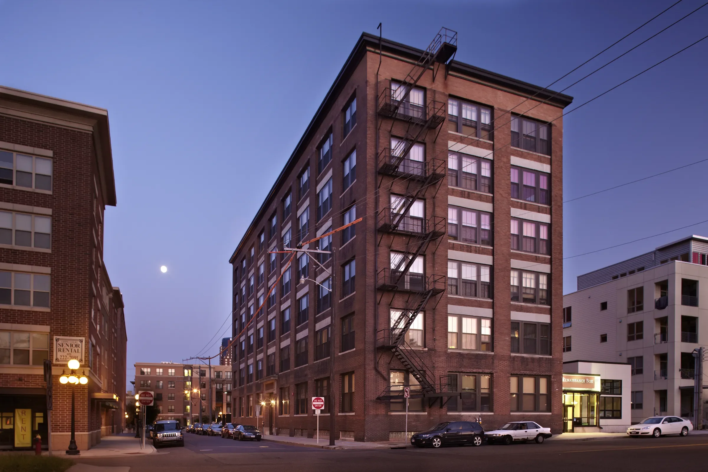A multi-story brick residential building with a fire escape on the side, situated on a city street at dusk with parked cars, streetlights, and neighboring buildings.