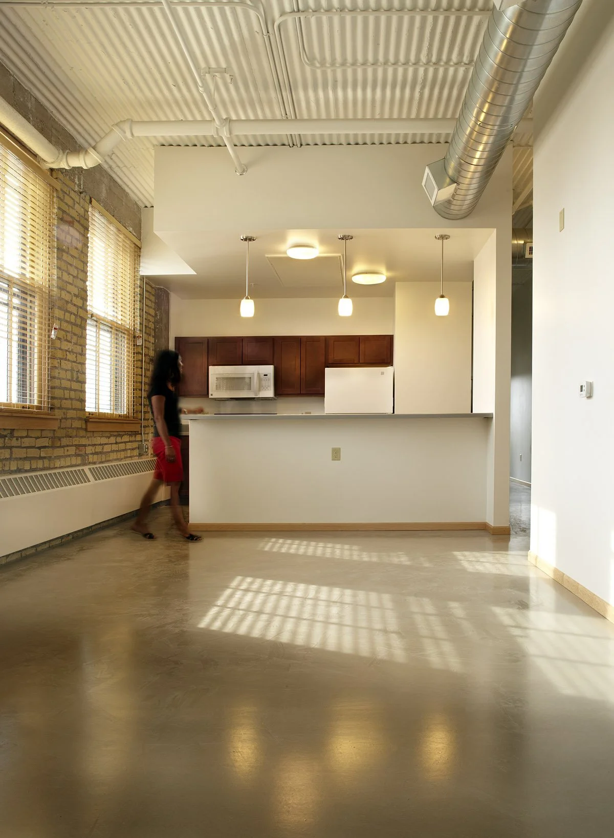 A woman walking in an open-concept kitchen with brick walls and large windows, with sunlight creating shadows on the floor.