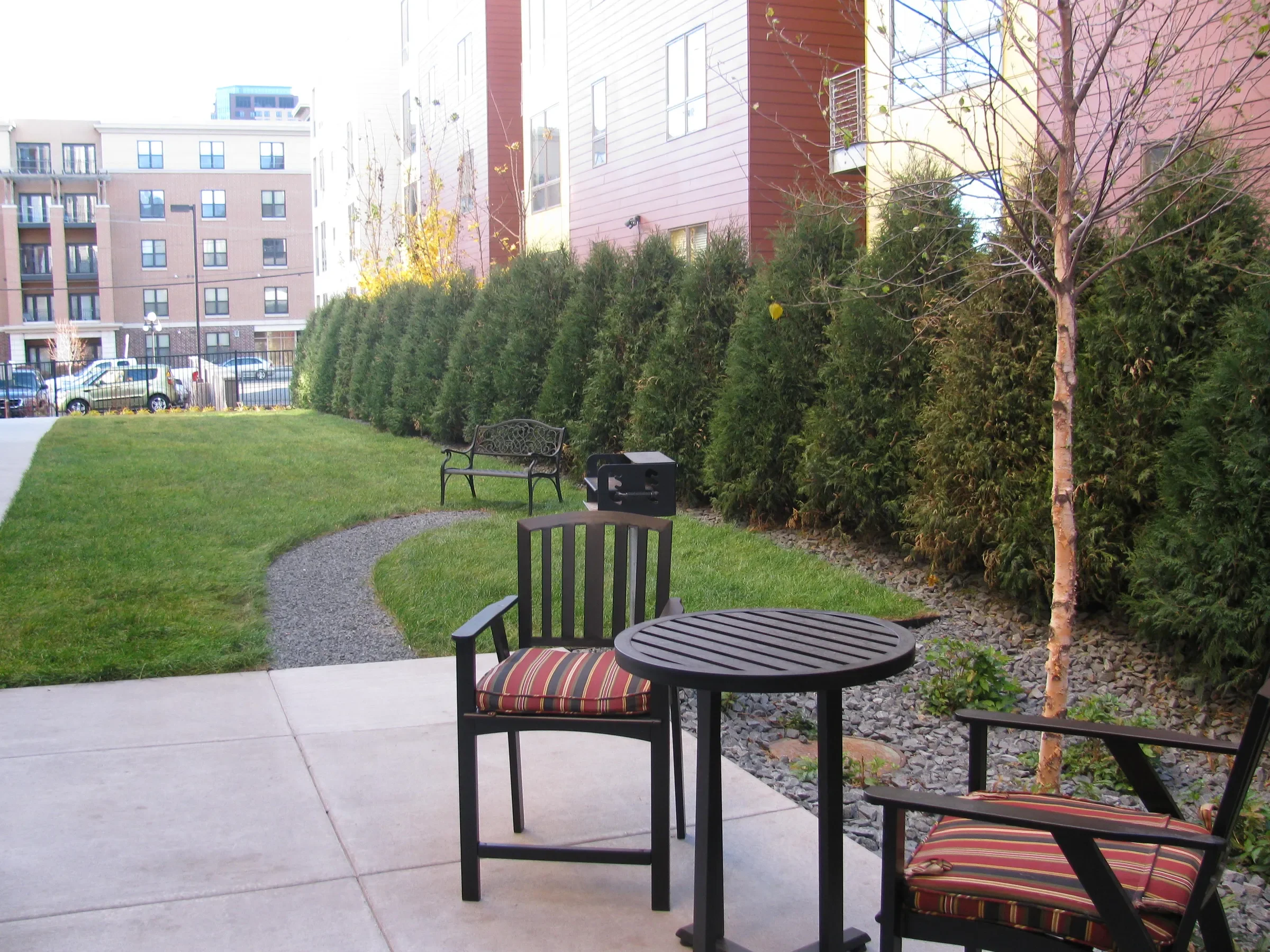 A small outdoor patio with black metal furniture, including a round table and two chairs with striped cushions. Behind it, a landscaped yard with a curved gravel pathway, green grass, a bench, and a row of tall bushes with an apartment building in th
