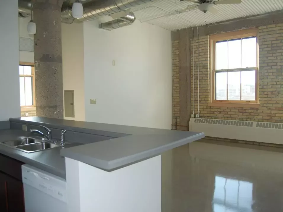 Empty industrial-style apartment with exposed brick, window, and kitchen counter with sink.