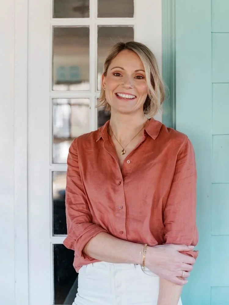 A woman with short blonde hair, wearing a rust-colored button-up shirt and white pants, standing outside a building with a glass door and turquoise siding, smiling at the camera.