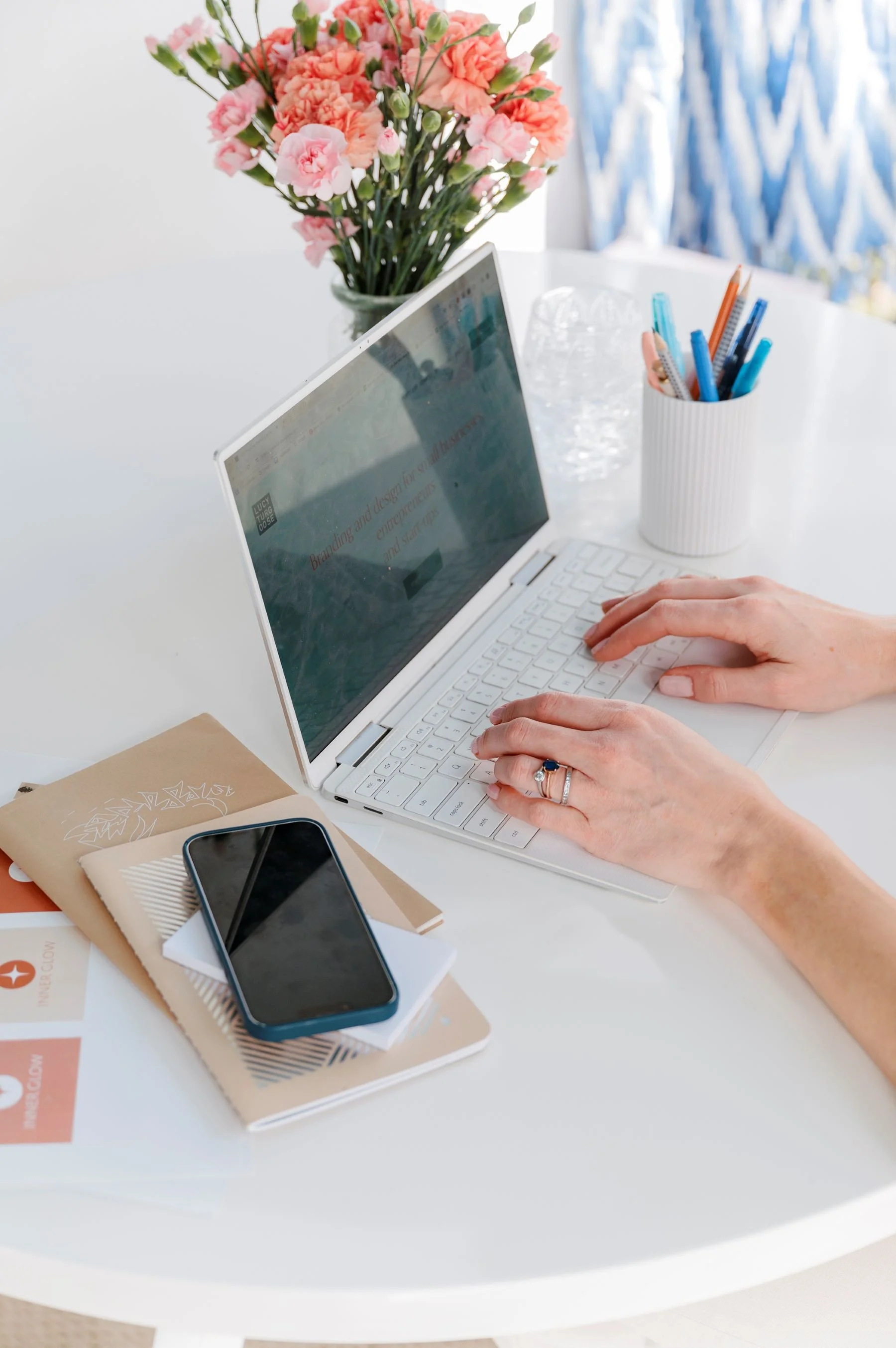A person's hands typing on a white laptop at a white table, with a vase of pink carnations and a container of pens nearby, and a smartphone and notebooks on the table.