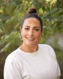 A woman with dark hair in a bun, wearing hoop earrings and a light-colored top, smiling outdoors with greenery in the background.